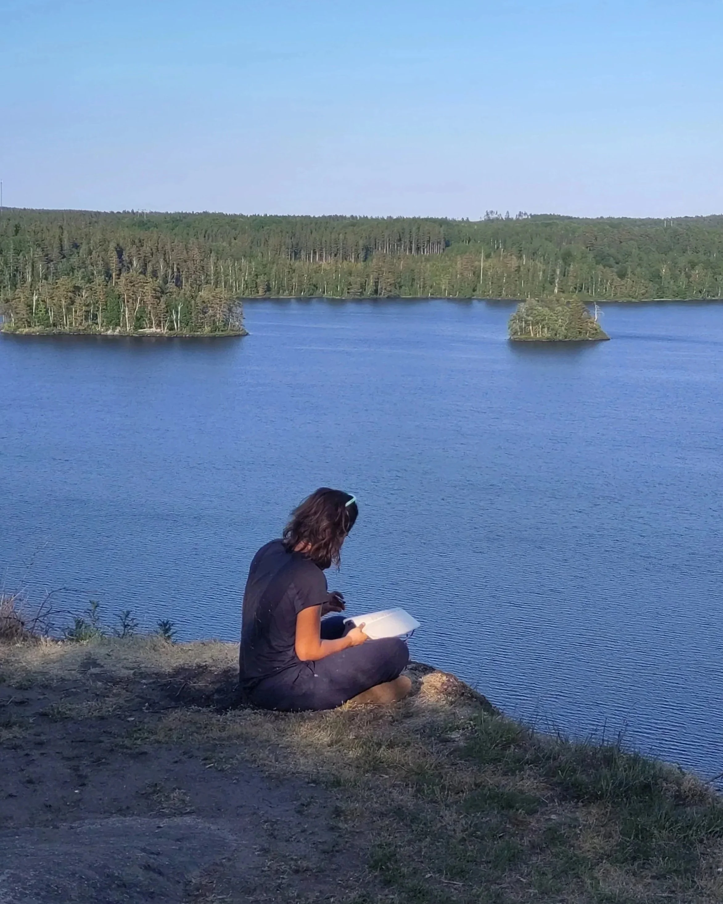 Une femme assise sur un rivage, regardant son carnet près d'un lac entouré d'arbres et de petites îles, sous un ciel clair.