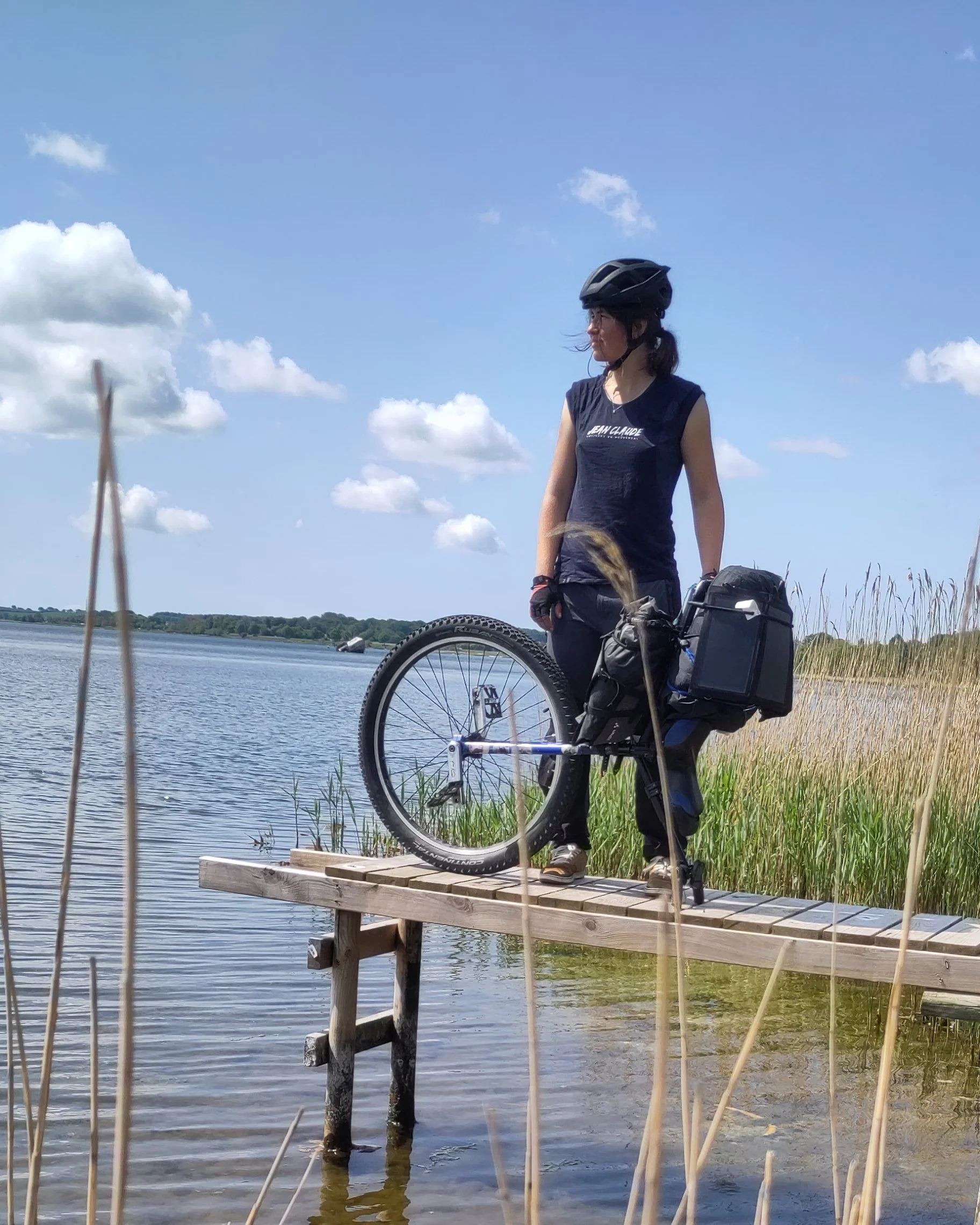Femme avec un casque de vélo, portant un sac à dos, debout sur une petite plateforme en bois au bord d'une rivière, avec un monocycle à côté d'elle, sous un ciel bleu avec quelques nuages.
