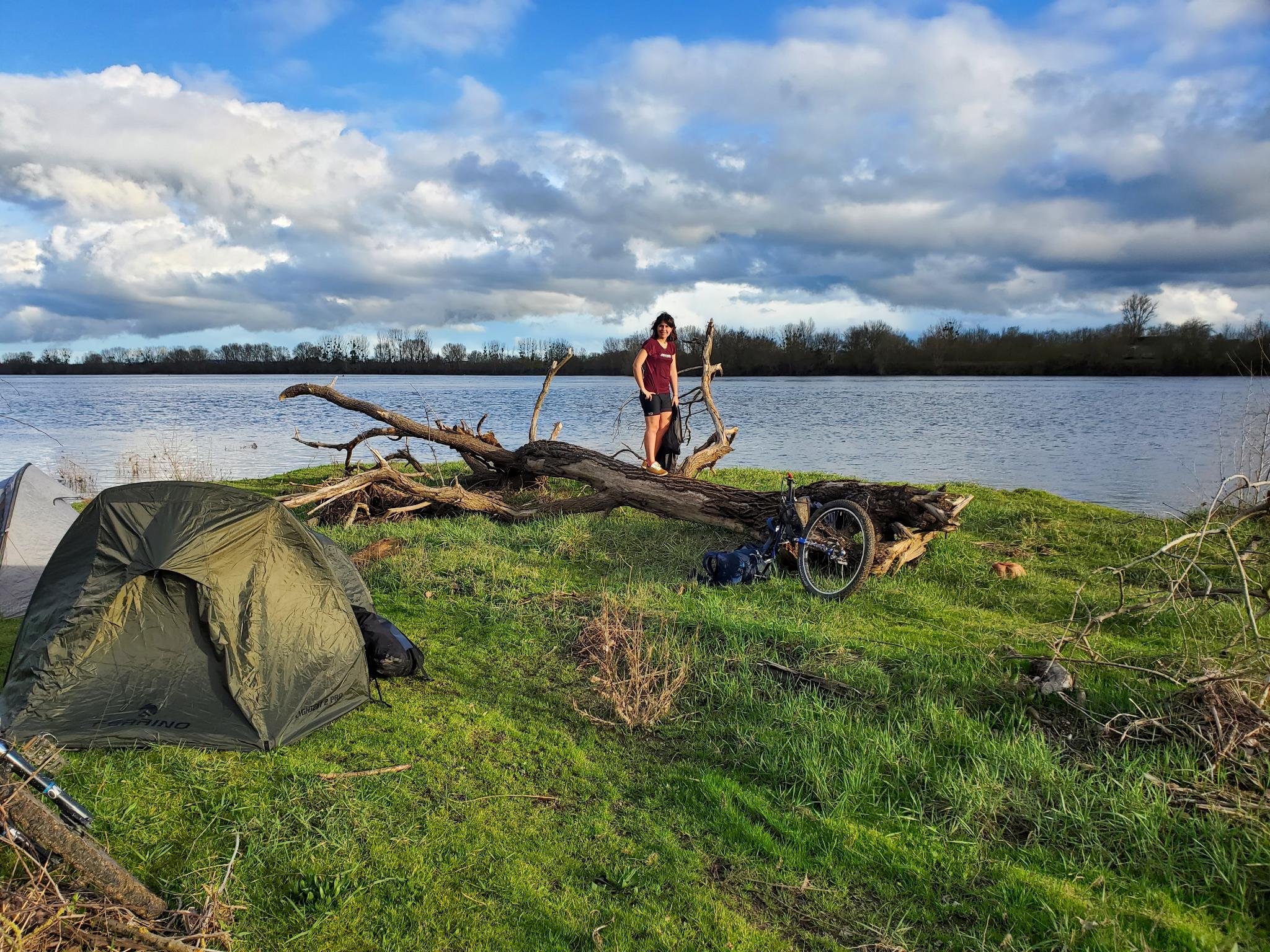 Une femme se tient sur un tronc d'arbre tombé près d'un lac, avec un monocycle à ses côtés, sous un ciel nuageux, autour de tentes installées sur l'herbe.