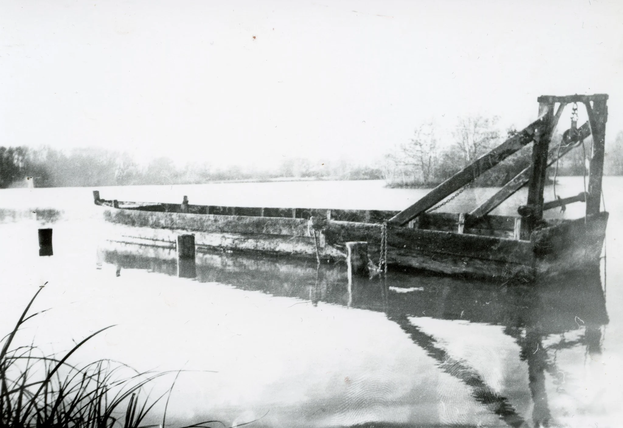 Photographie en noir et blanc d'un bateau en bois abandonné dans l'eau, avec un paysage naturel en arrière-plan.