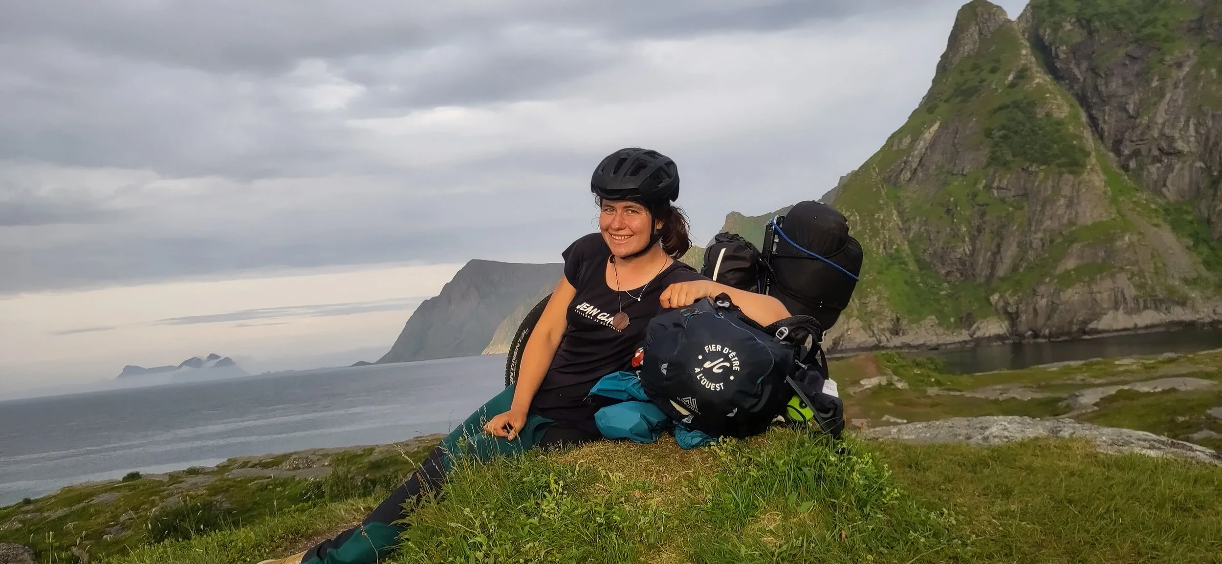 Femme souriante portant un casque de vélo, assise sur l'herbe avec son sac à dos et son monocycle au bord d'une falaise, paysage de montagnes et de mer derrière elle en Norvège.