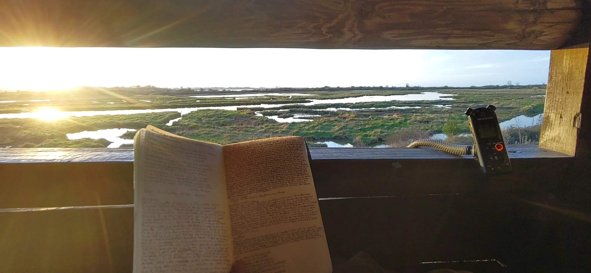 Vue d'un paysage de marais avec des eaux calmes sous un ciel clair, prise depuis une cabane en bois avec un livre, un enregistreur, sur une fenêtre ouverte au coucher du soleil.
