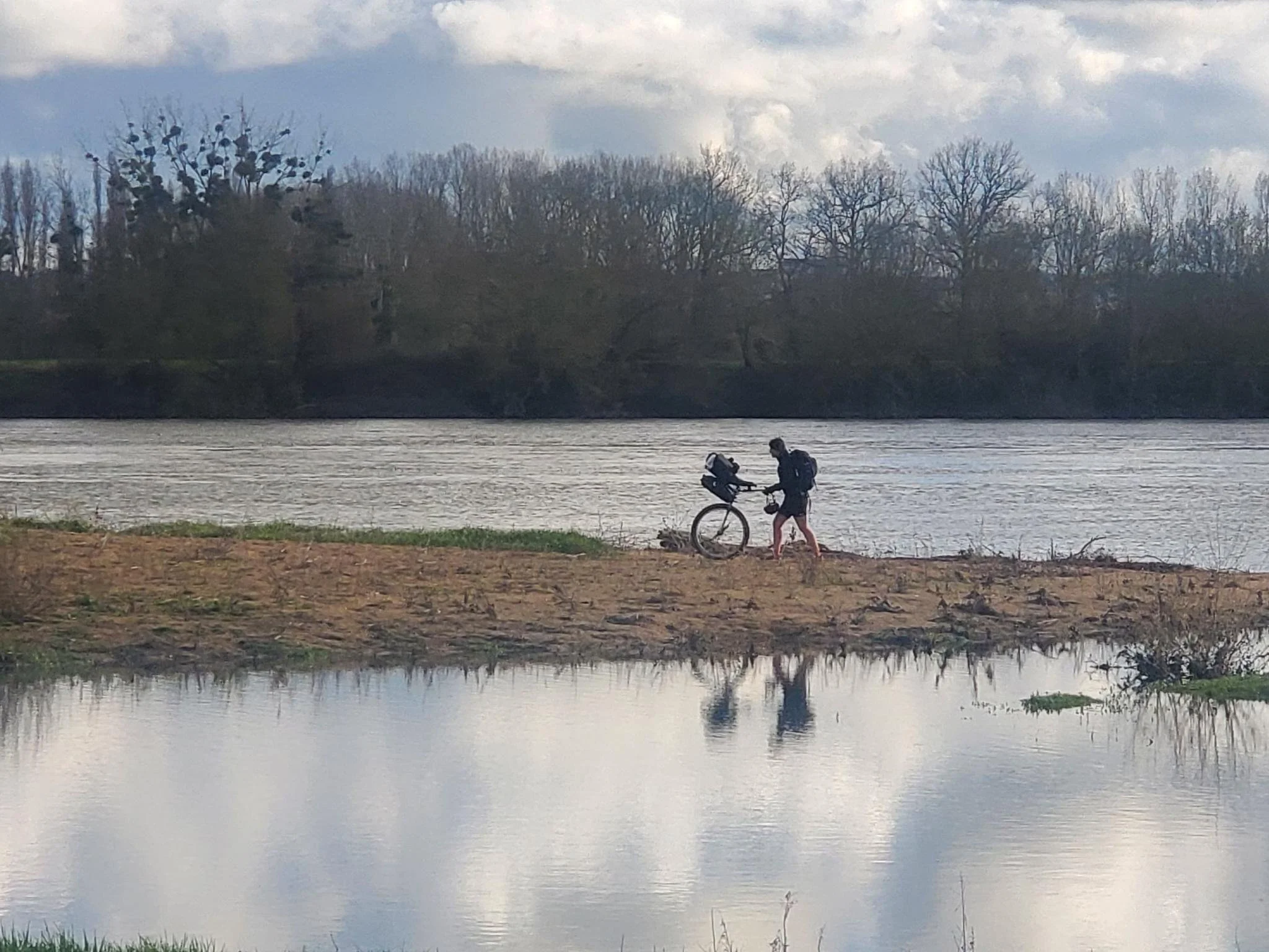 Une personne avec un monocycle et un sac à dos marchant sur la berge d'une rivière, avec des arbres au fond et un ciel nuageux.
