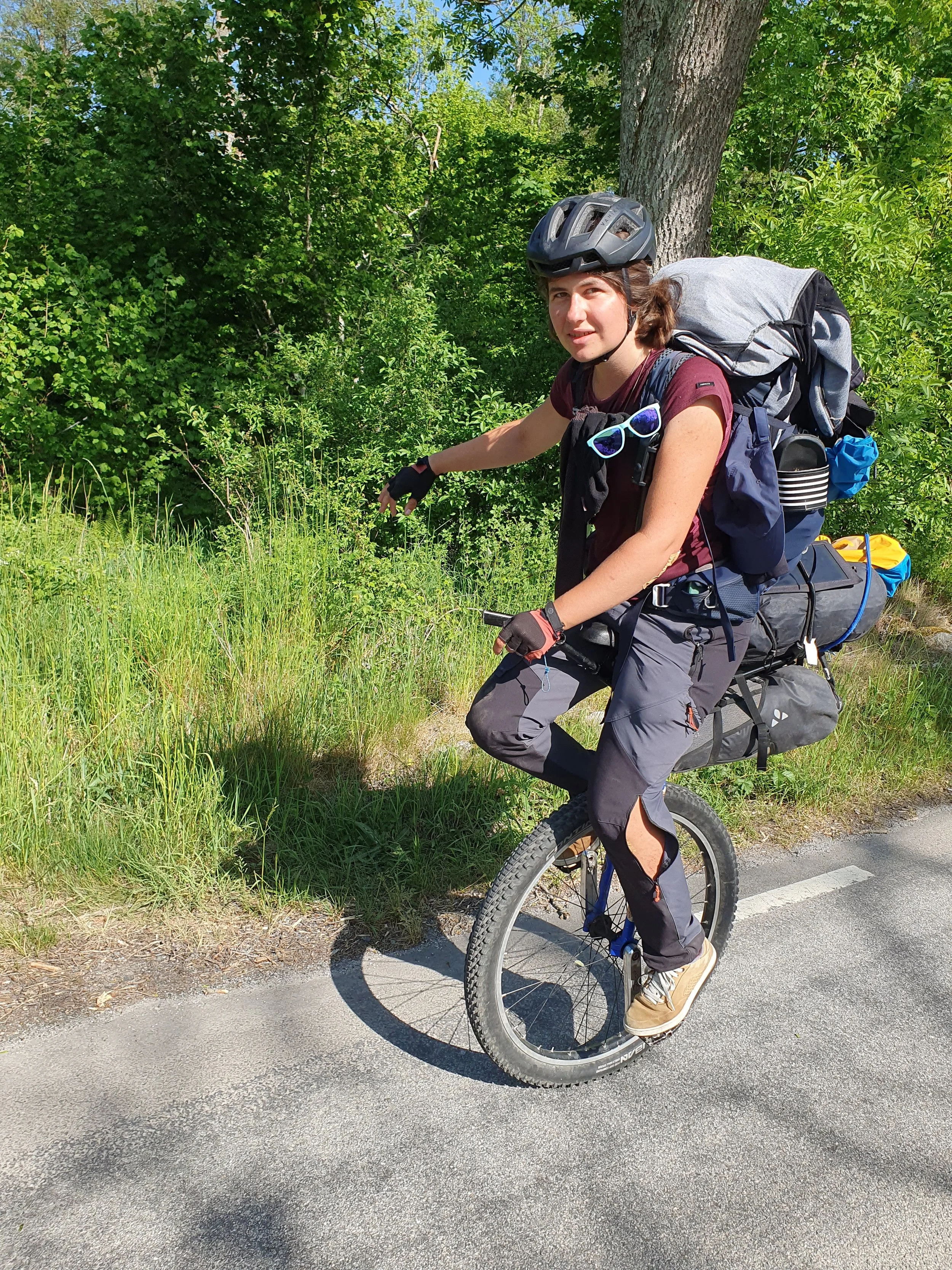 Femme en monocycle avec casque, sac à dos, lunettes de soleil, vêtements de randonnée, pointant vers la végétation à côté d’un arbre sur une route en plein air, par temps ensoleillé.