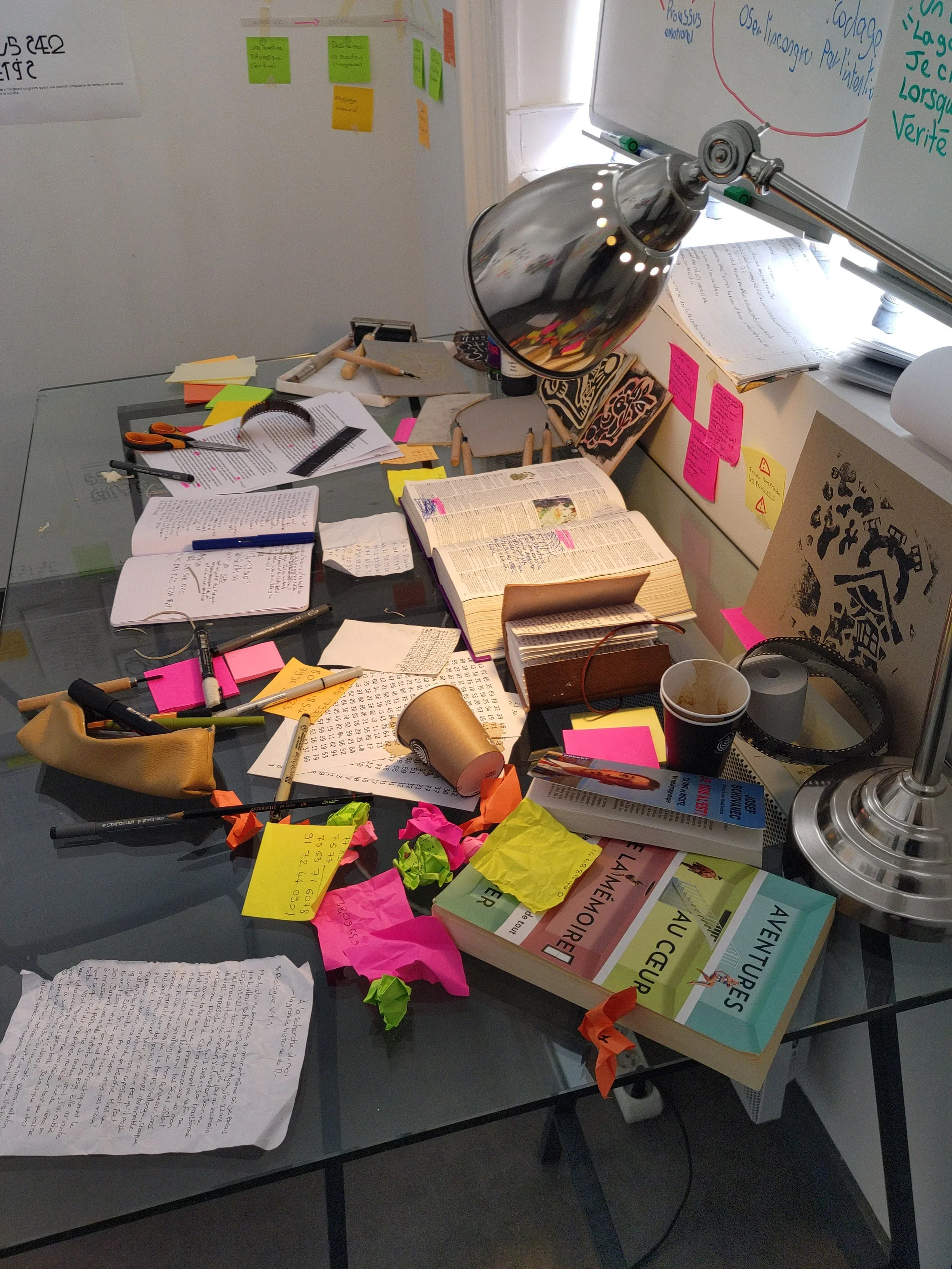 Bureau désordonné avec des livres, des notes autocollantes, des papiers, une lampe de bureau, des marqueurs, une tasse vide, une paire de ciseaux, et un tableau blanc avec des écrits, suggérant un espace de travail ou d'étude chaotique.