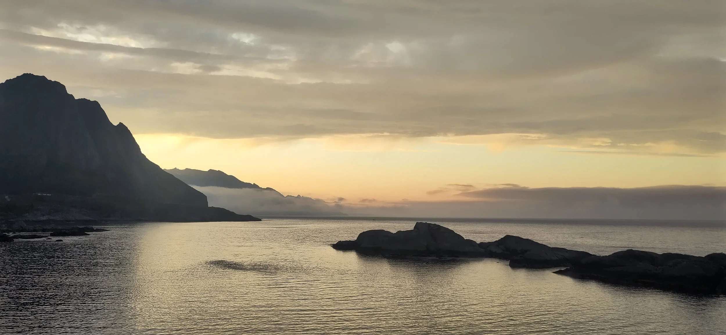 Côte rocheuse au coucher du soleil avec montagnes en arrière-plan et mer calme.