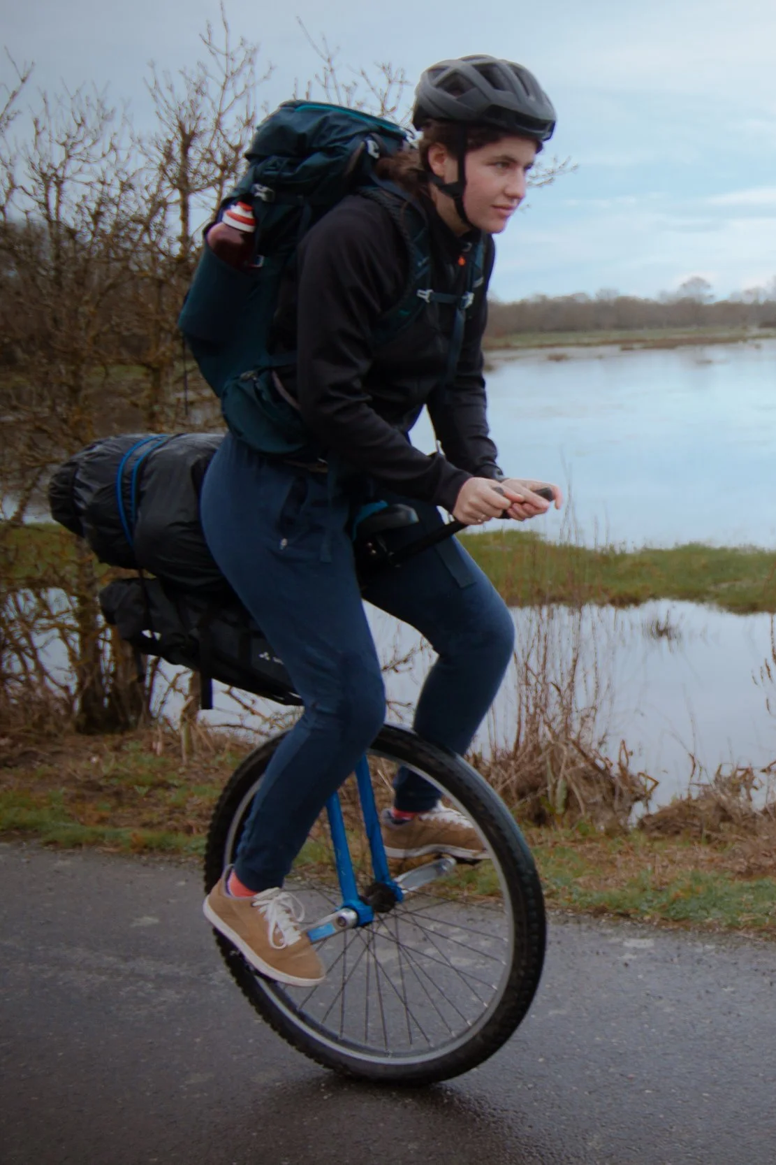 Jeune femme à vélo sur un chemin près d'un lac, portant un casque et un sac à dos, par temps nuageux.