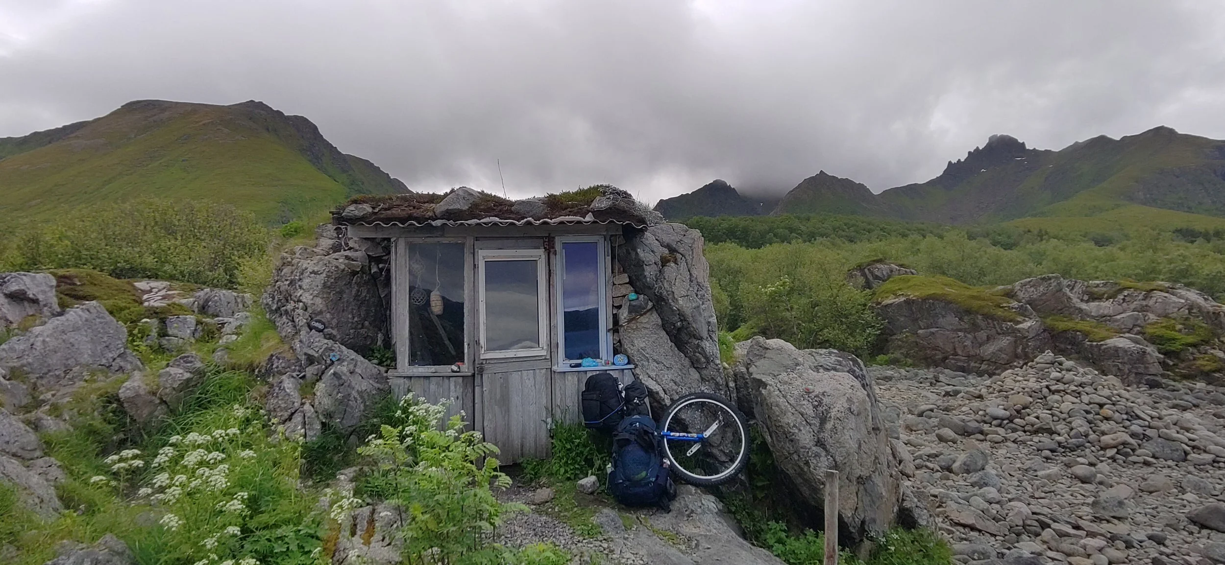 Petite cabane en pierre et bois construite contre un rocher, avec des montagnes verdoyantes en arrière-plan, sous un ciel nuageux. Il y a un monocycle, un sac à dos et une petite fenêtre devant la cabane.