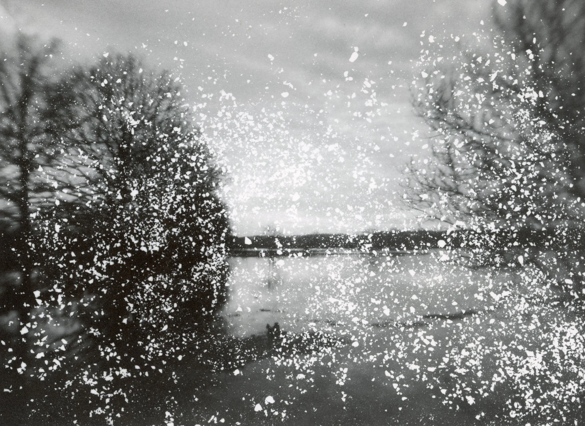 Une vue d'un lac avec des arbres sans feuilles de chaque côté, vue à travers une fenêtre sale ou givrée, avec des particules ou des flocons de neige en premier plan, en noir et blanc.