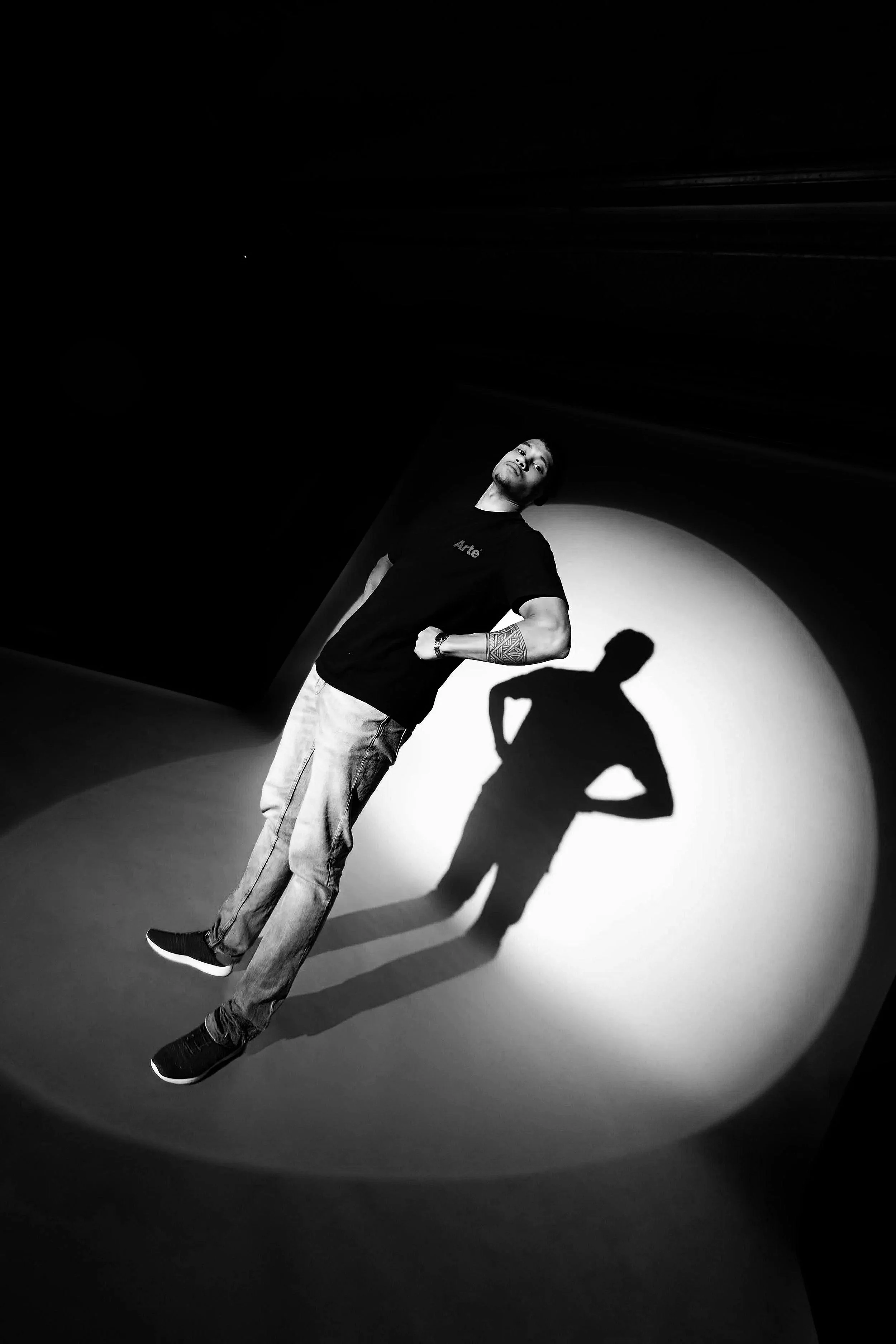 Man poseert in een donkere omgeving met een schijnwerper die zijn schaduw projecteert op de muur achter hem, alles in zwart-wit.