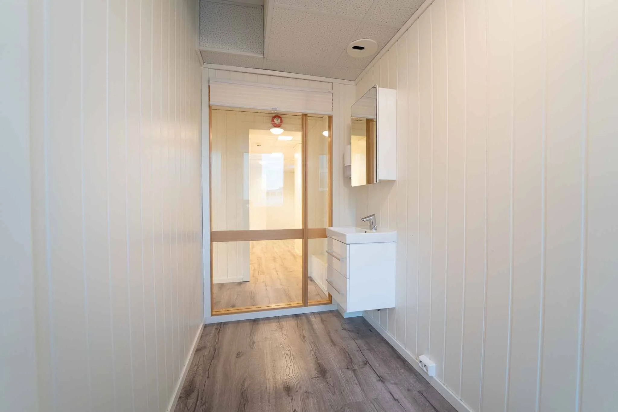 En liten renovert vaskebutikk med en hvit vask og speil på veggen, adskilt av en glassdør til et annet rom.