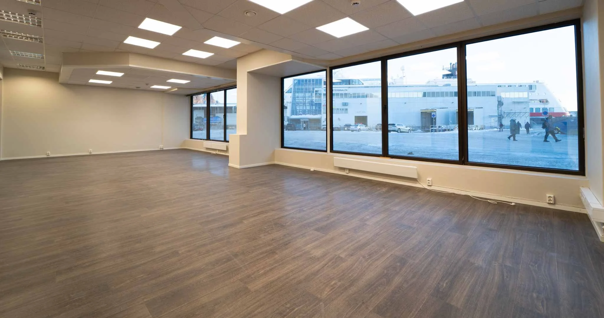 Tomt og moderne rom med store vinduer mot gaten, hvite vegger og mørkt tremønster gulv.