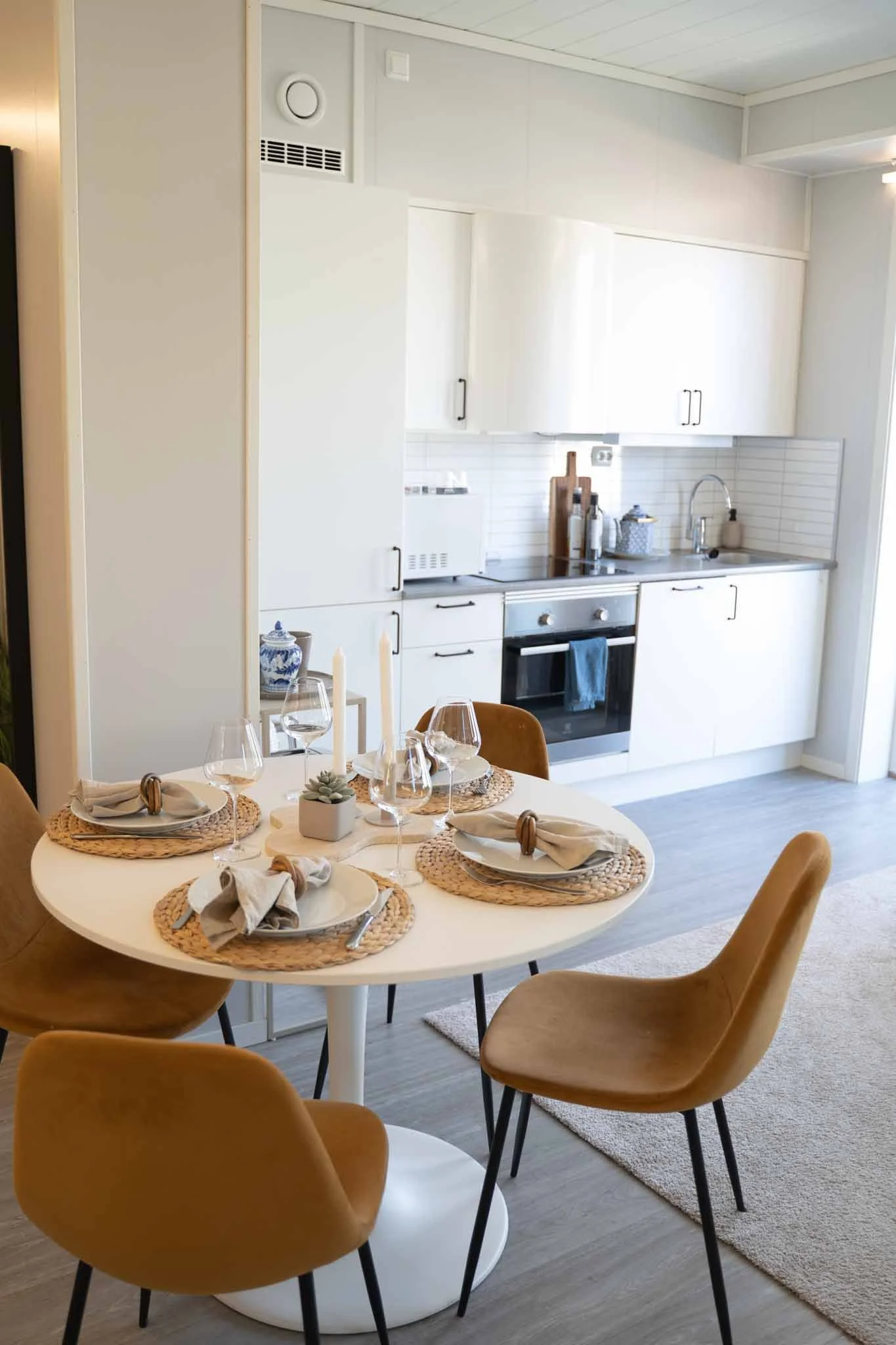 A round dining table set for four, with placemats, napkins, wine glasses, and a small centerpiece, next to a white kitchen with cabinets, an oven, and a sink.
