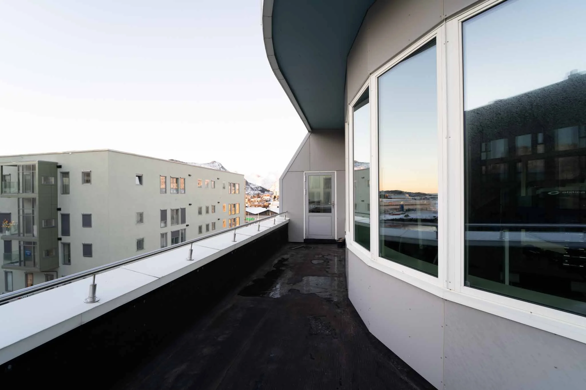 Balkong på en moderne bygning med store vinduer, utsikt over nærliggende leilighetsbygninger og snødekte fjell i bakgrunnen.