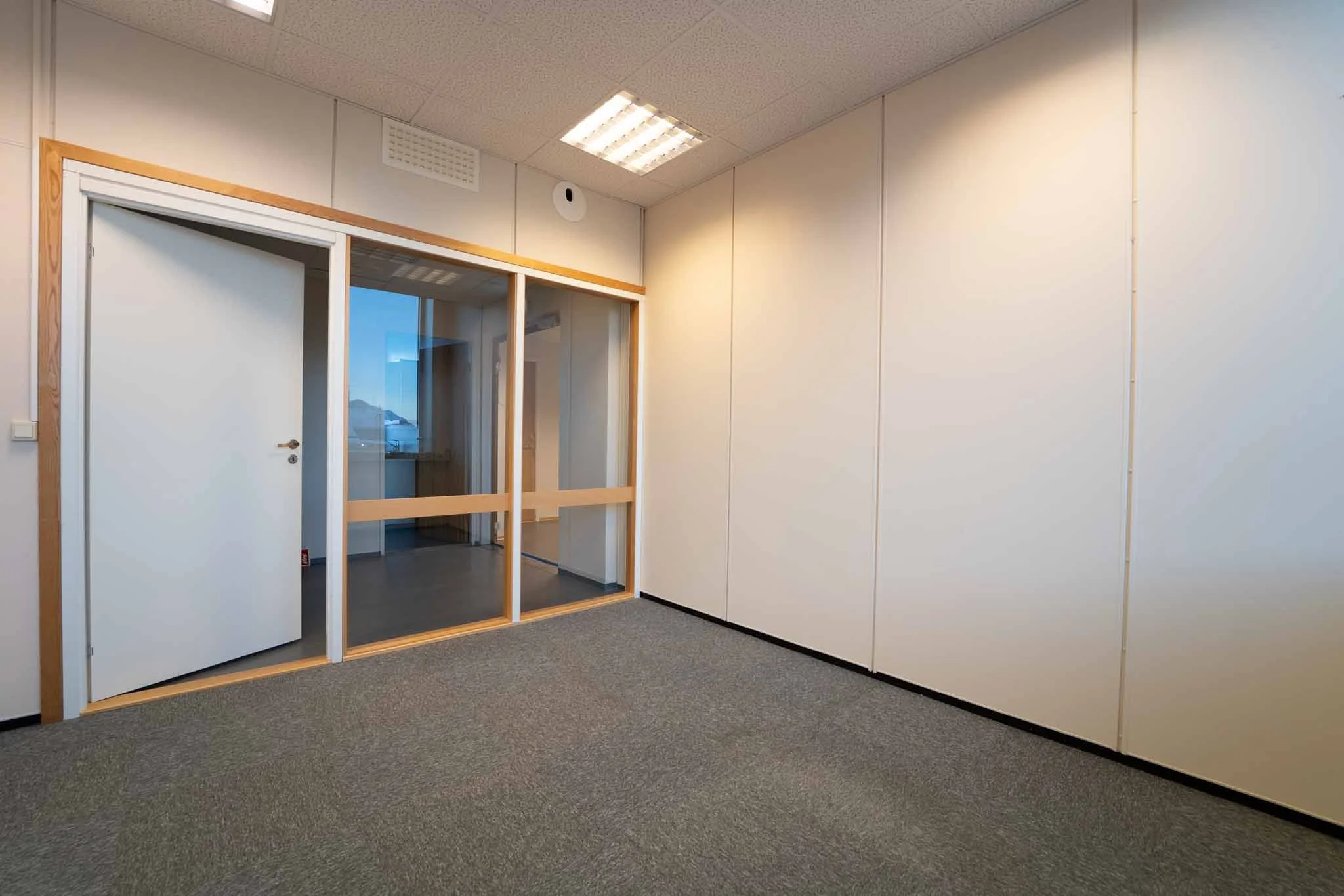 Tomt kontor med hvite vegger, grå teppe og glassdør som leder til et annet rom.