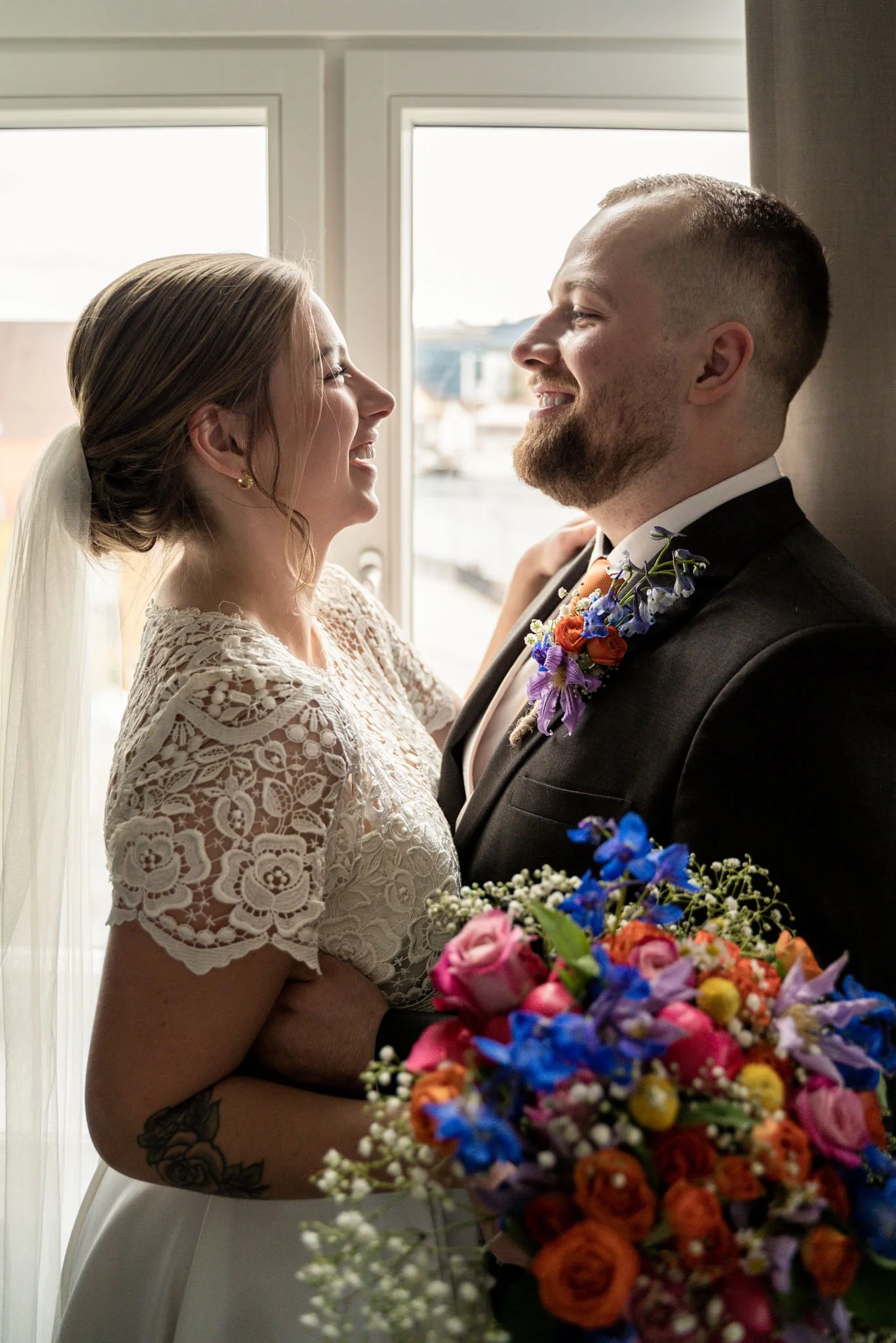 Et brudepar står ansikt til ansikt, smiler og ser på hverandre, foran et vindu med utsikt over byen, med en fargerik blomsterbukett i forgrunnen.