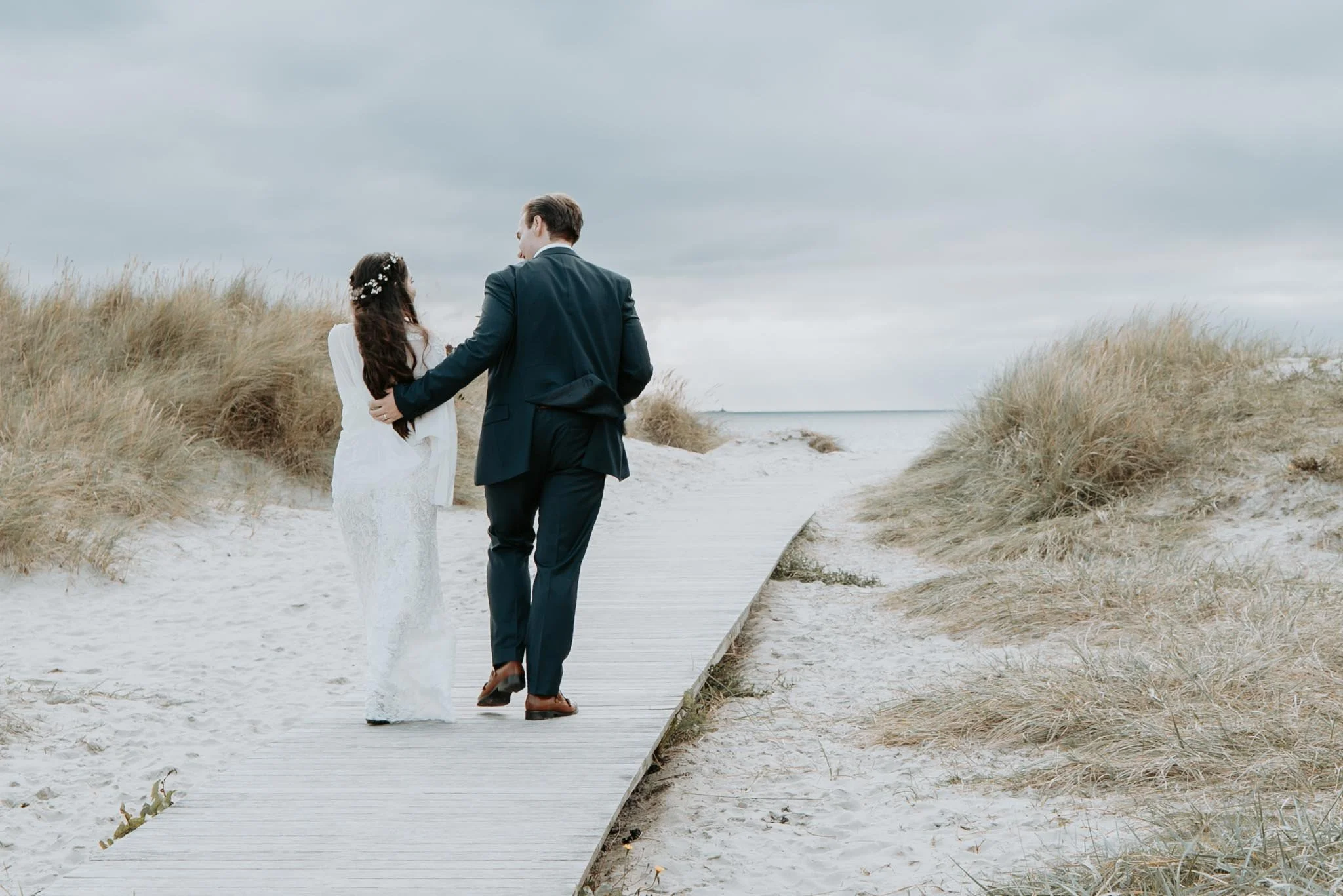 Et ungt par i bryllup kledd i formelle klær, går hånd i hånd langs en strandpromenade med sanddyner og kystlandskap under en overskyet himmel.
