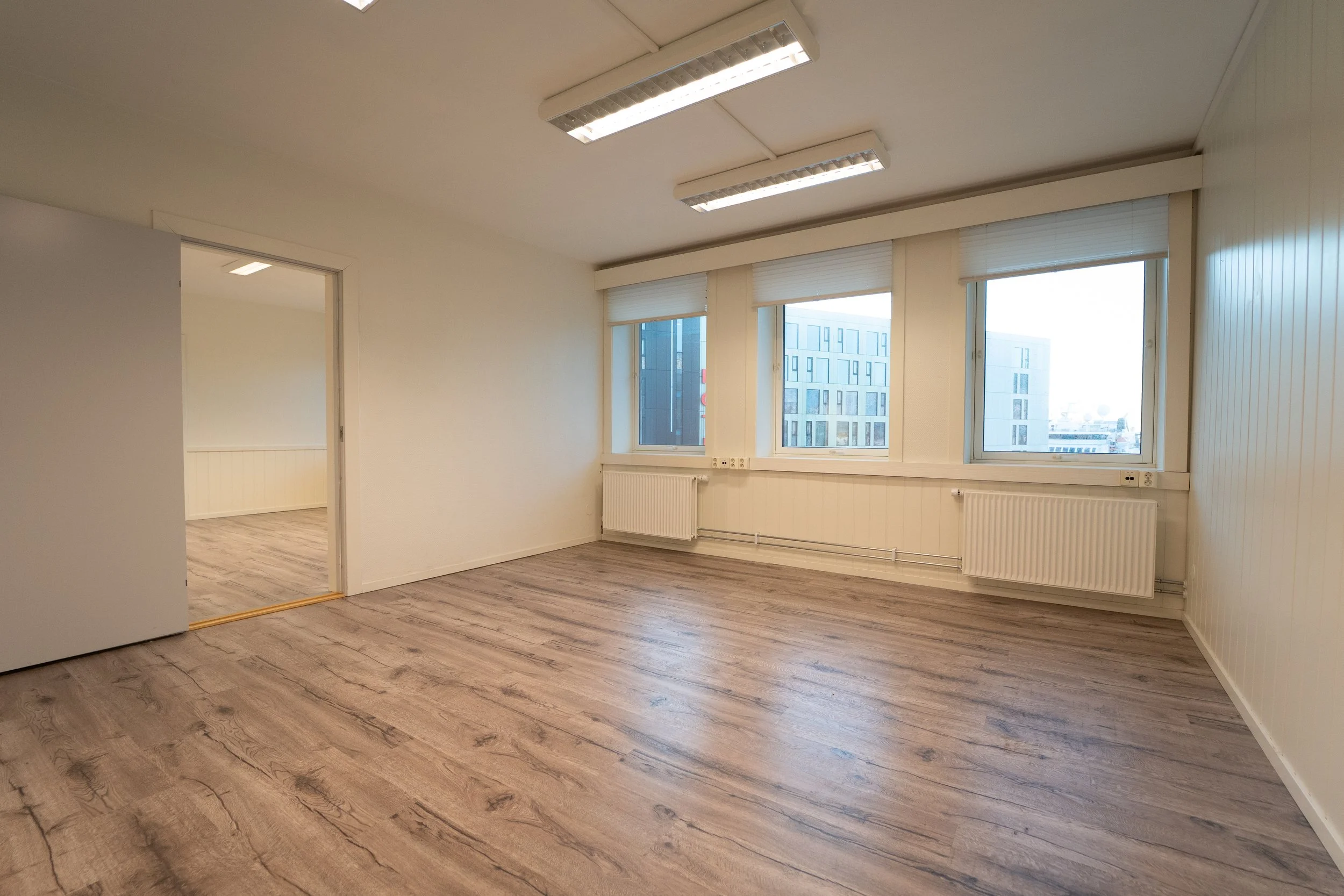 Tomt rom med store vinduer, lyst treparkett og hvite vegger.