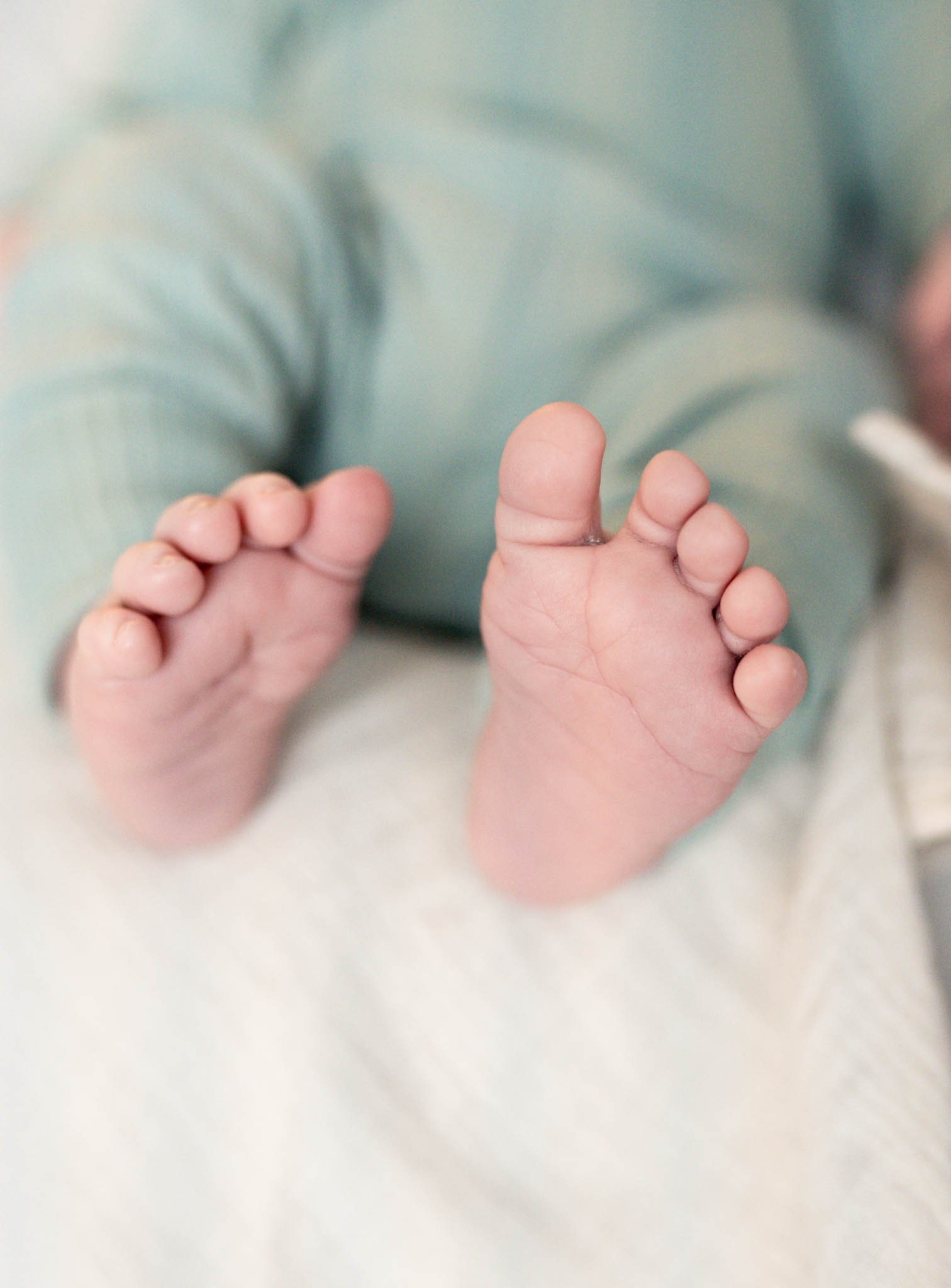 Nærbilder av små babyfingre som holder hverandre, med en bakgrunn som er utusikkert, trolig en person iført en lyseblå eller grønn pyjamas.