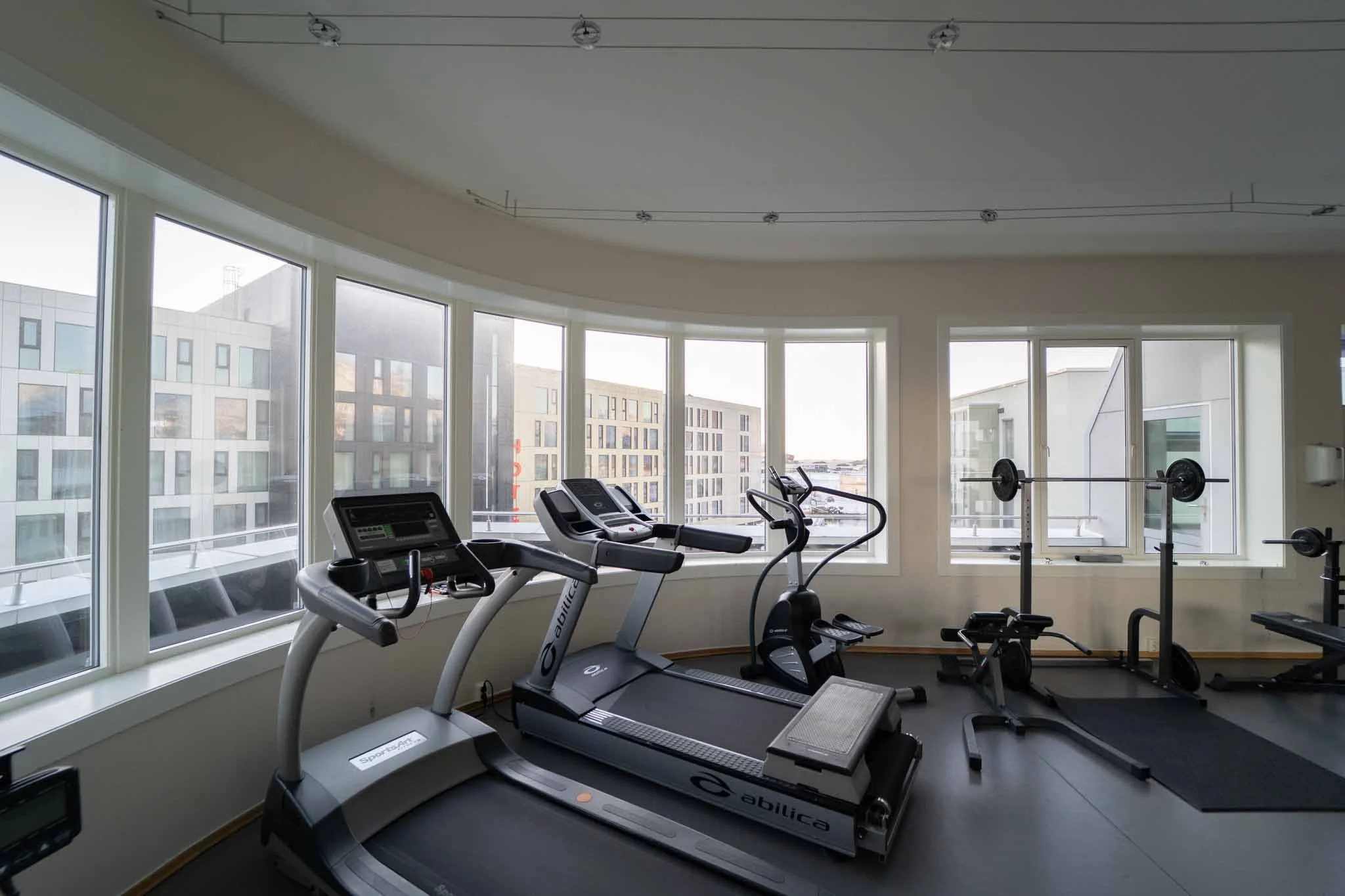 Fitnessrom med tredemøller, sykkel og vektstang ved store vinduer med utsikt over byen