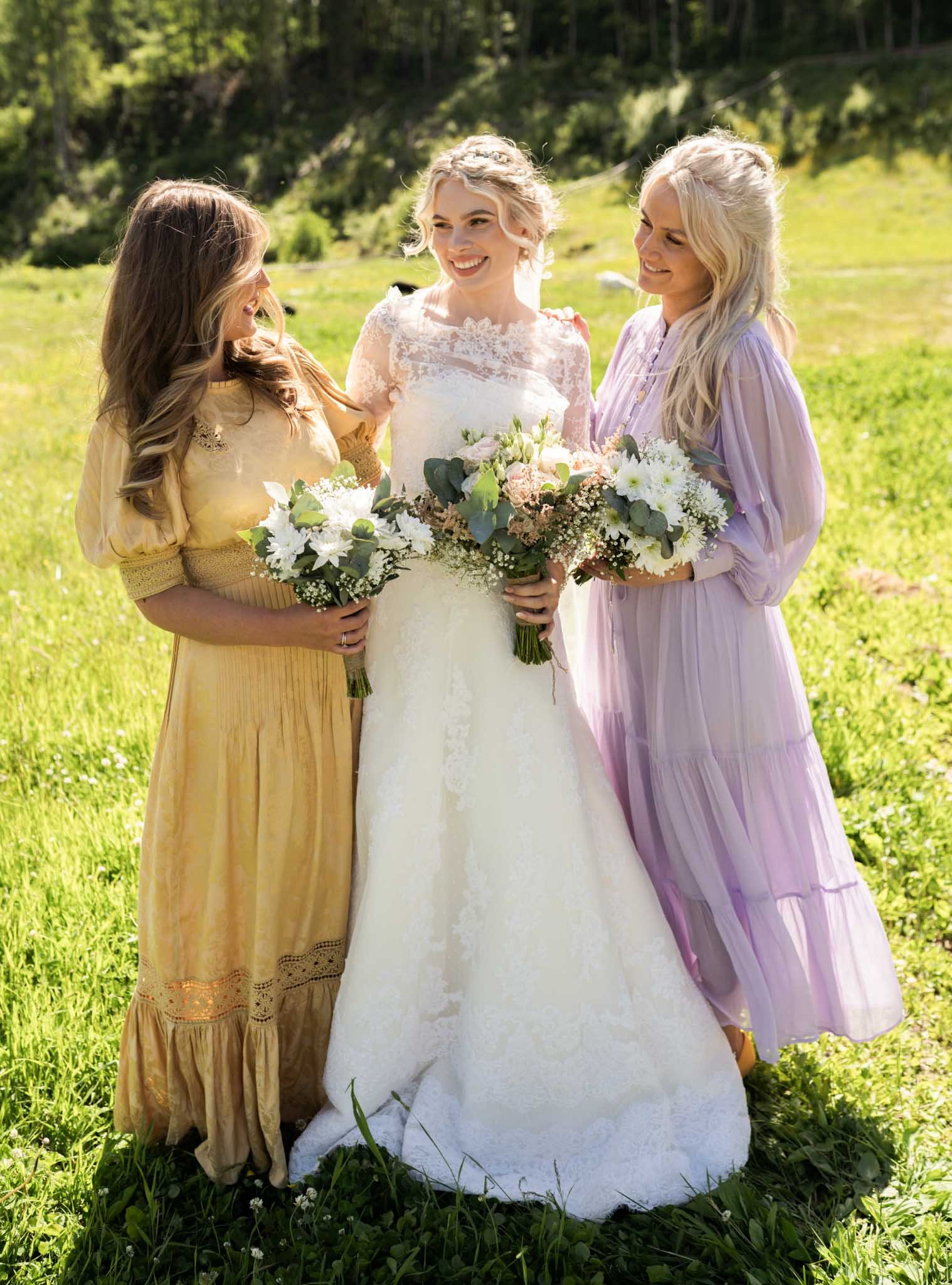 En brud i en hvit kjole holder en bukett blomster, omkranset av to kvinner i elegante kjoler, på en grønn eng med trær i bakgrunnen.