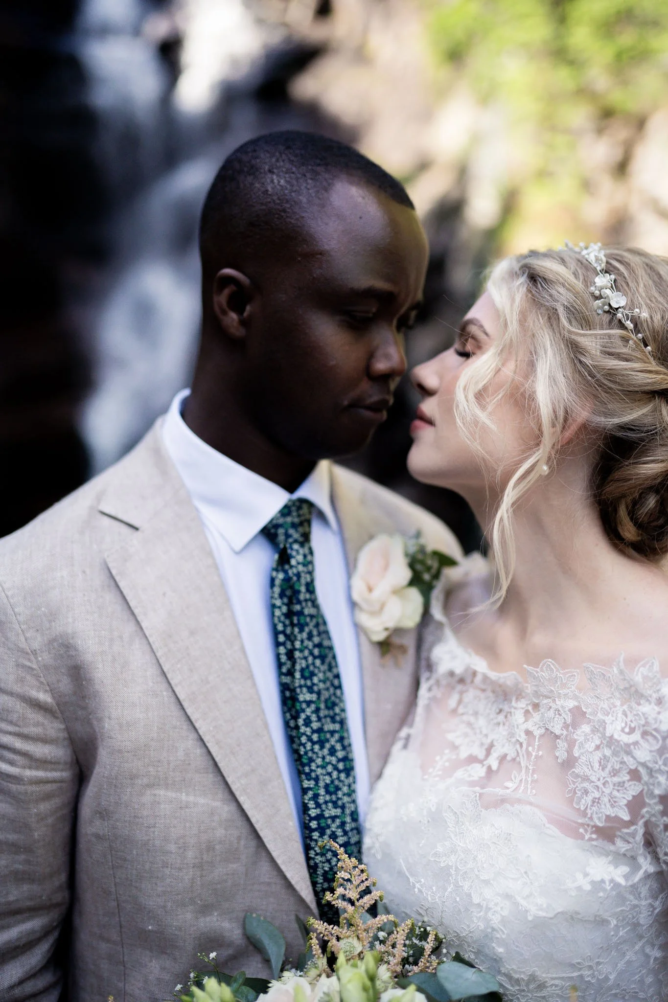 Nærbilde av et bryllupspar som står nært, med lukkede øyne, foran en naturlig bakgrunn. Mann i lys dress og hvit skjorte, kvinne i brudekjole med blonder og en blomsterkrans i håret, holder en bryllupskrans.