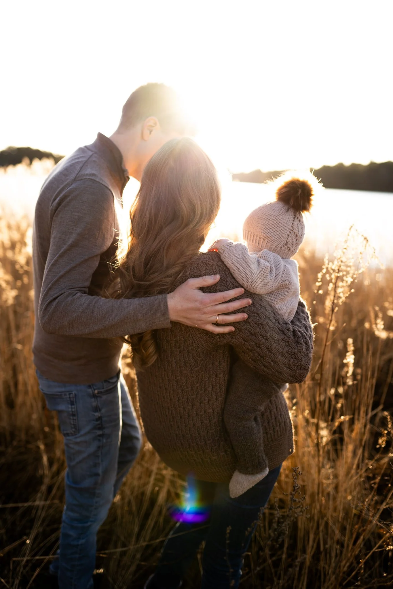 Golden hour. Mamma, pappa og en 6 måneder gammel jente står ved gylne strå i vannkanten.
