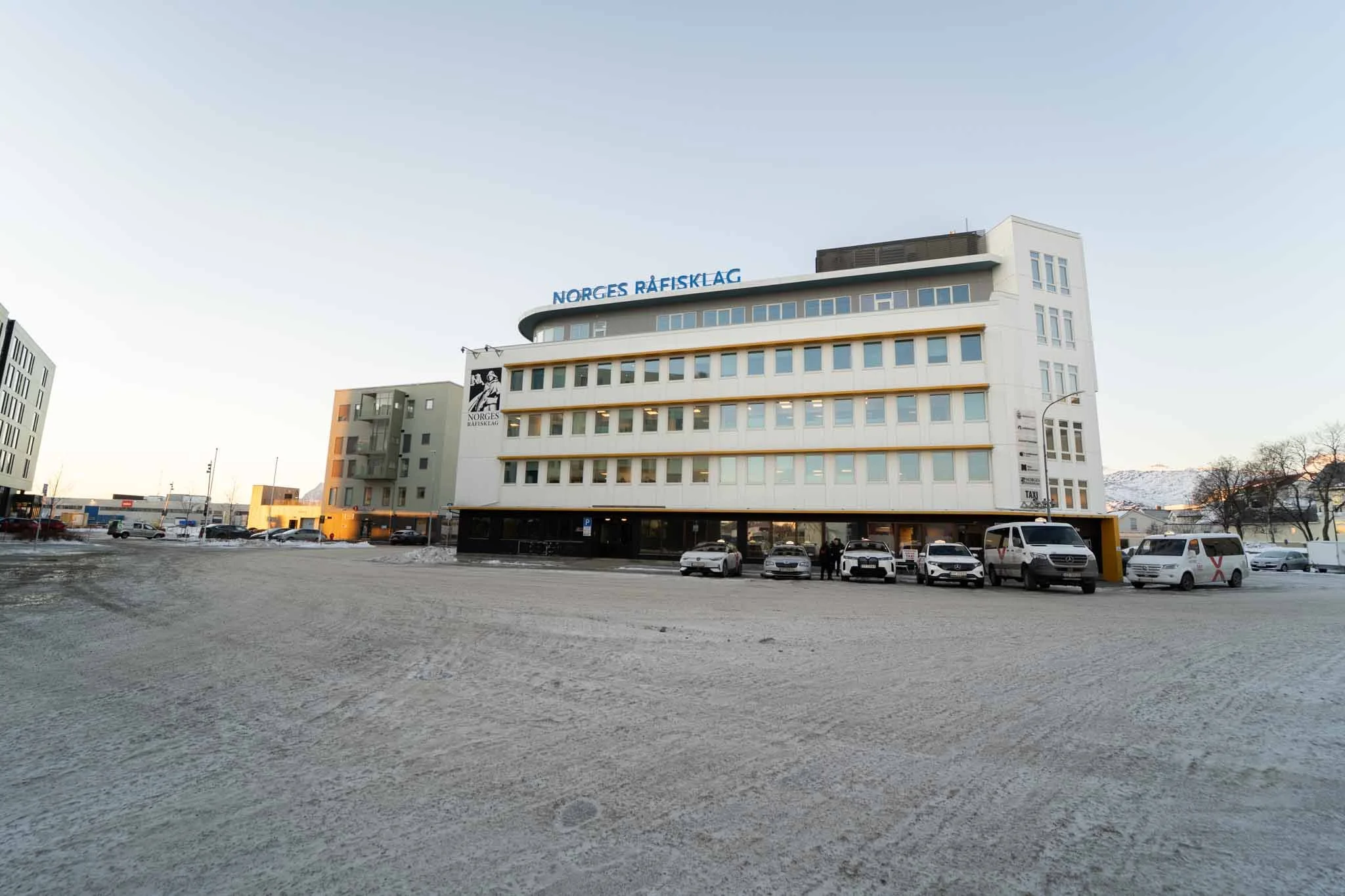 Et moderne kontorbygg med skiltet 'Norges Råfisklag' på taket, omgitt av et stort, snødekket parkeringsområde med flere biler og en lang rekke små bygninger mot en fjellbakgrunn under en klar himmel.