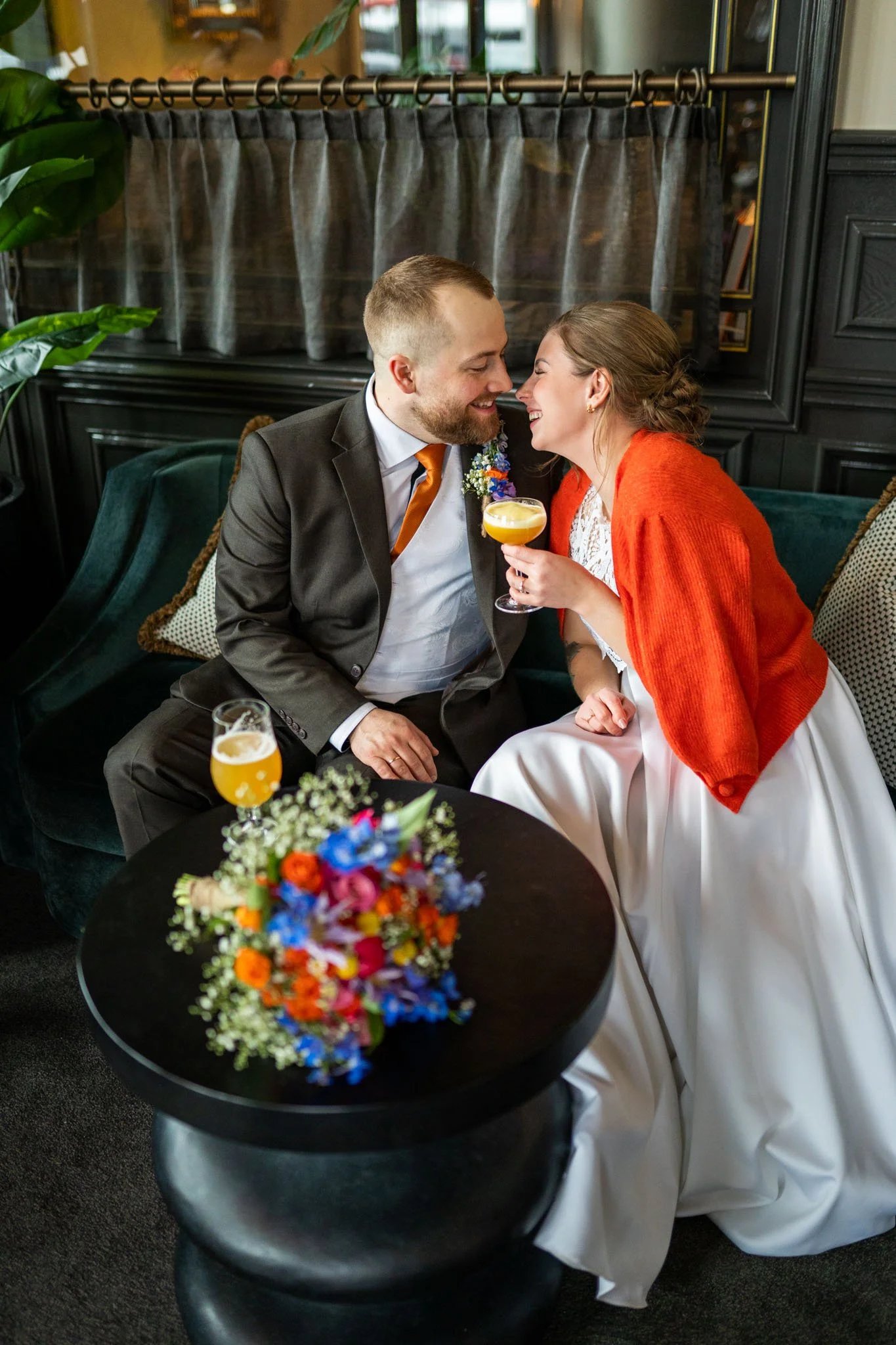 Et lykkelig par i bryllupsmote, mannen i dress med slips og bruden i hvit kjole med en oransje cardigan, imøtekommende og smilende, i en innendørs setting med blomster og dekorasjoner.