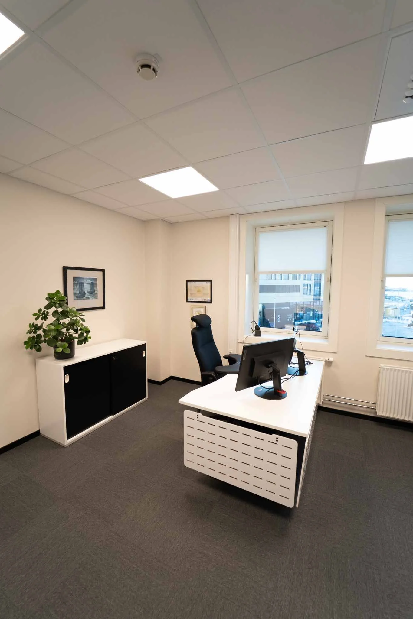 Et tomt kontor med skrivebord, kontorstol, dataskjerm, vinduer og en plante på en hvit sidebord.