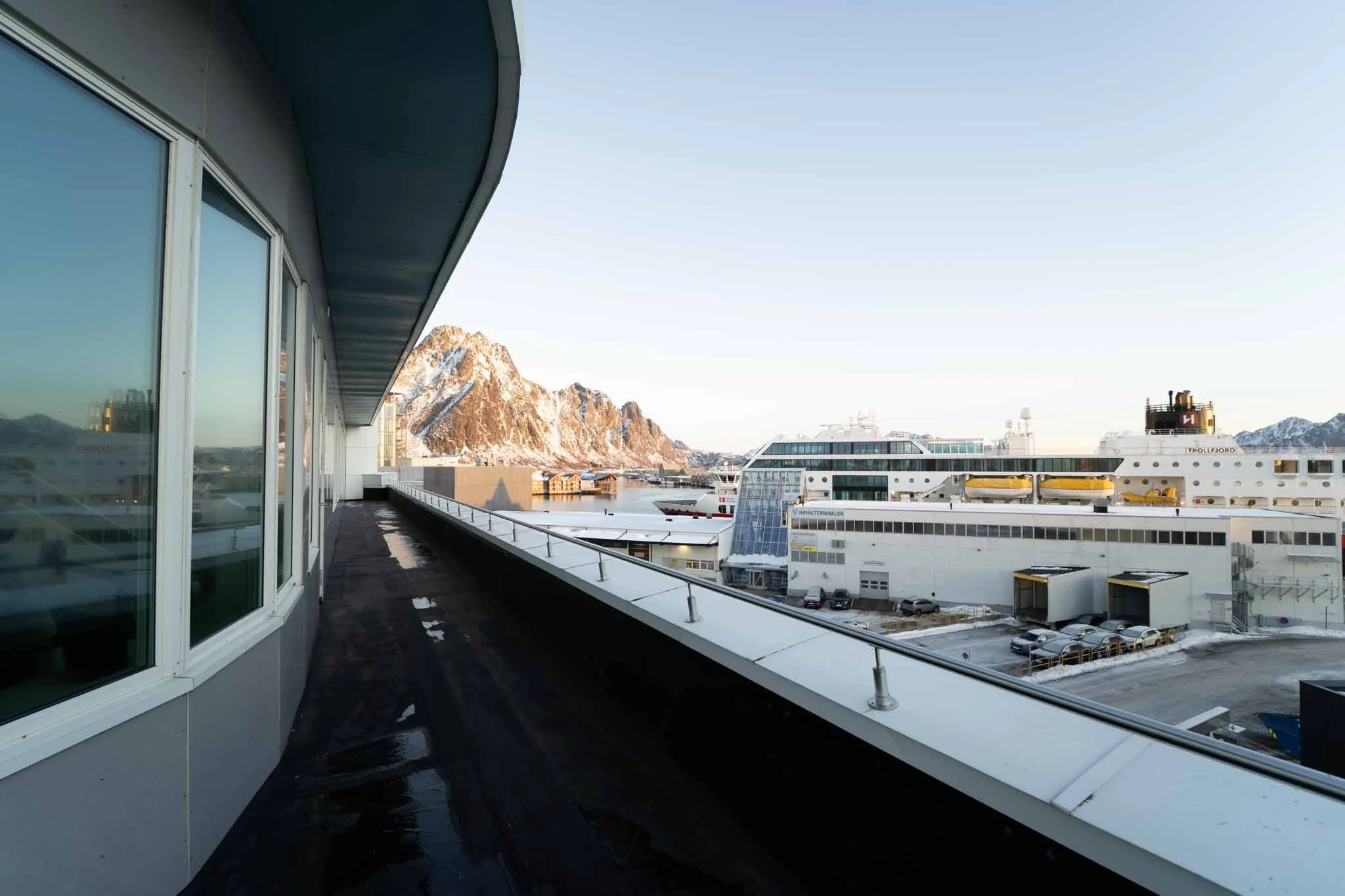 Utsikt fra en balkong på en cruiseskip med fjell i bakgrunnen, flere ferger og bygg under, klarvær, tidlig morgen eller sen ettermiddag.