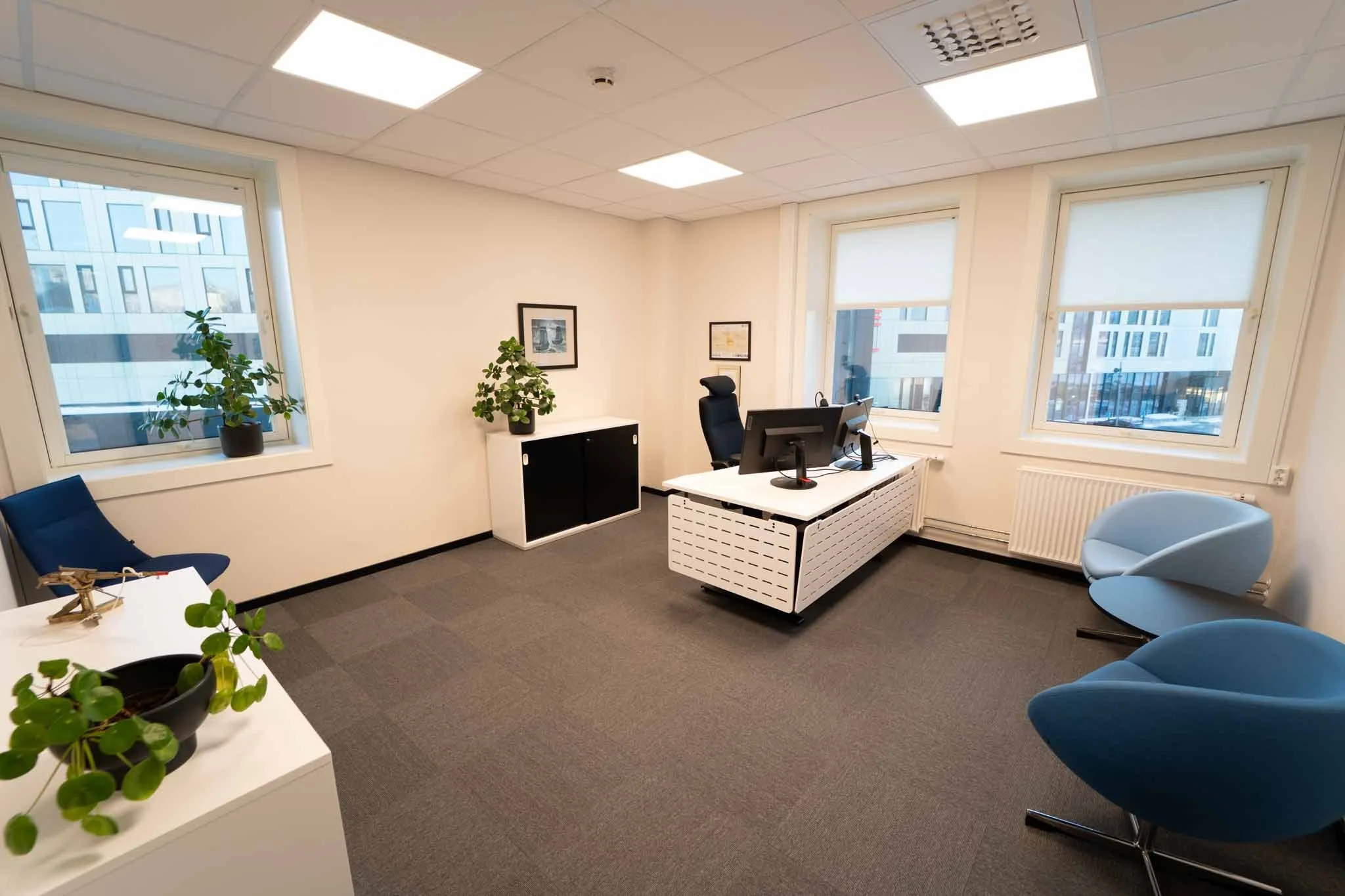 An office with a white desk, black office chair, two teal chairs, a white cabinet, and several potted plants next to large windows showing a cityscape.