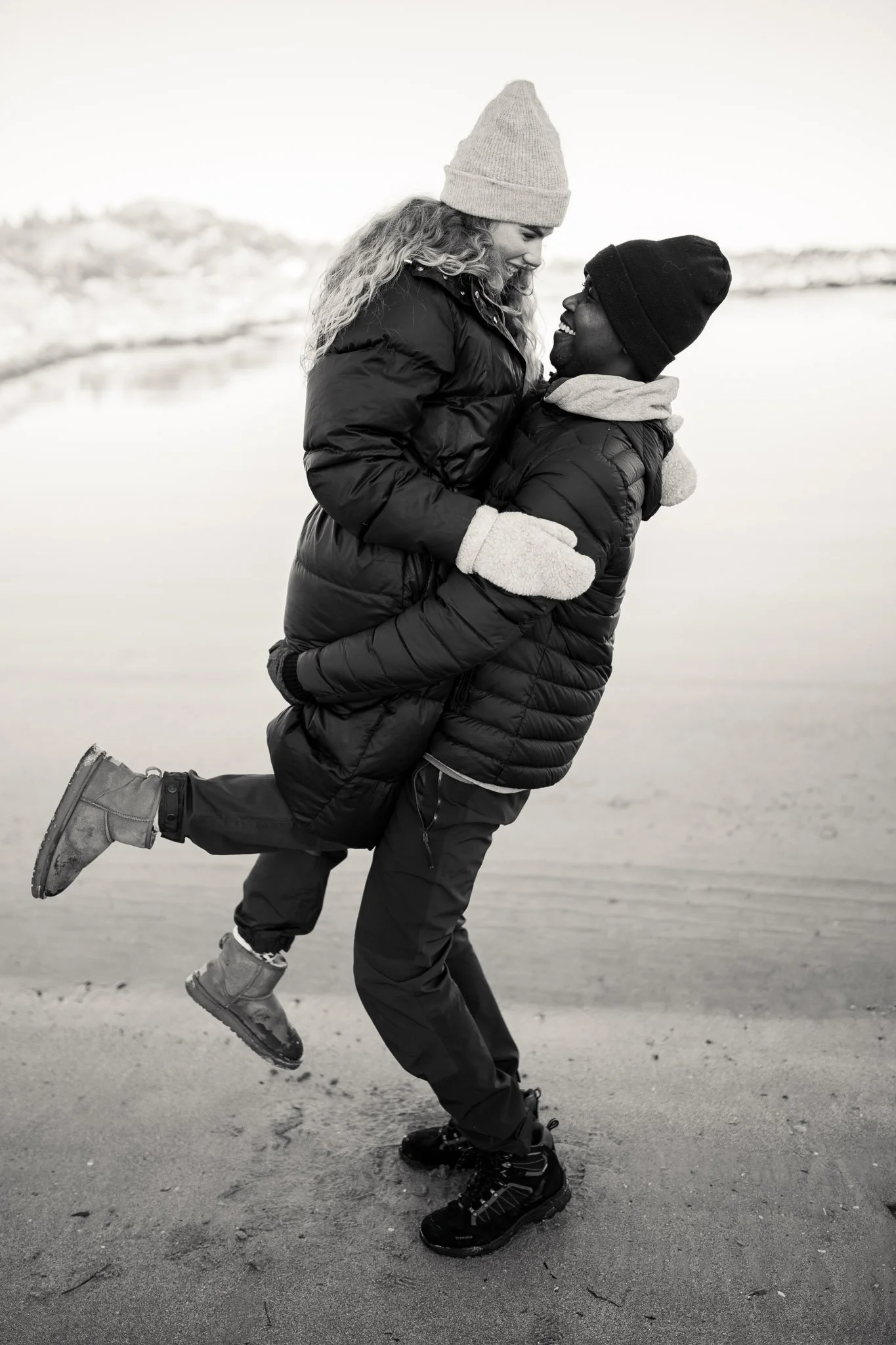 A black-and-white photo of a man and woman at a beach. The man is holding the woman off the ground, and they are smiling at each other. They are dressed warmly in winter jackets, hats, and gloves.