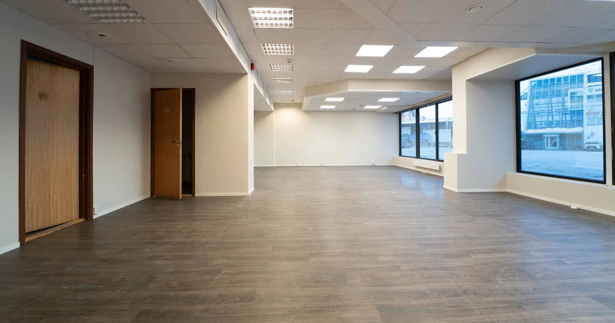 Tomt kontorlokale med store vinduer, lyse vegger og treparkettgulv.