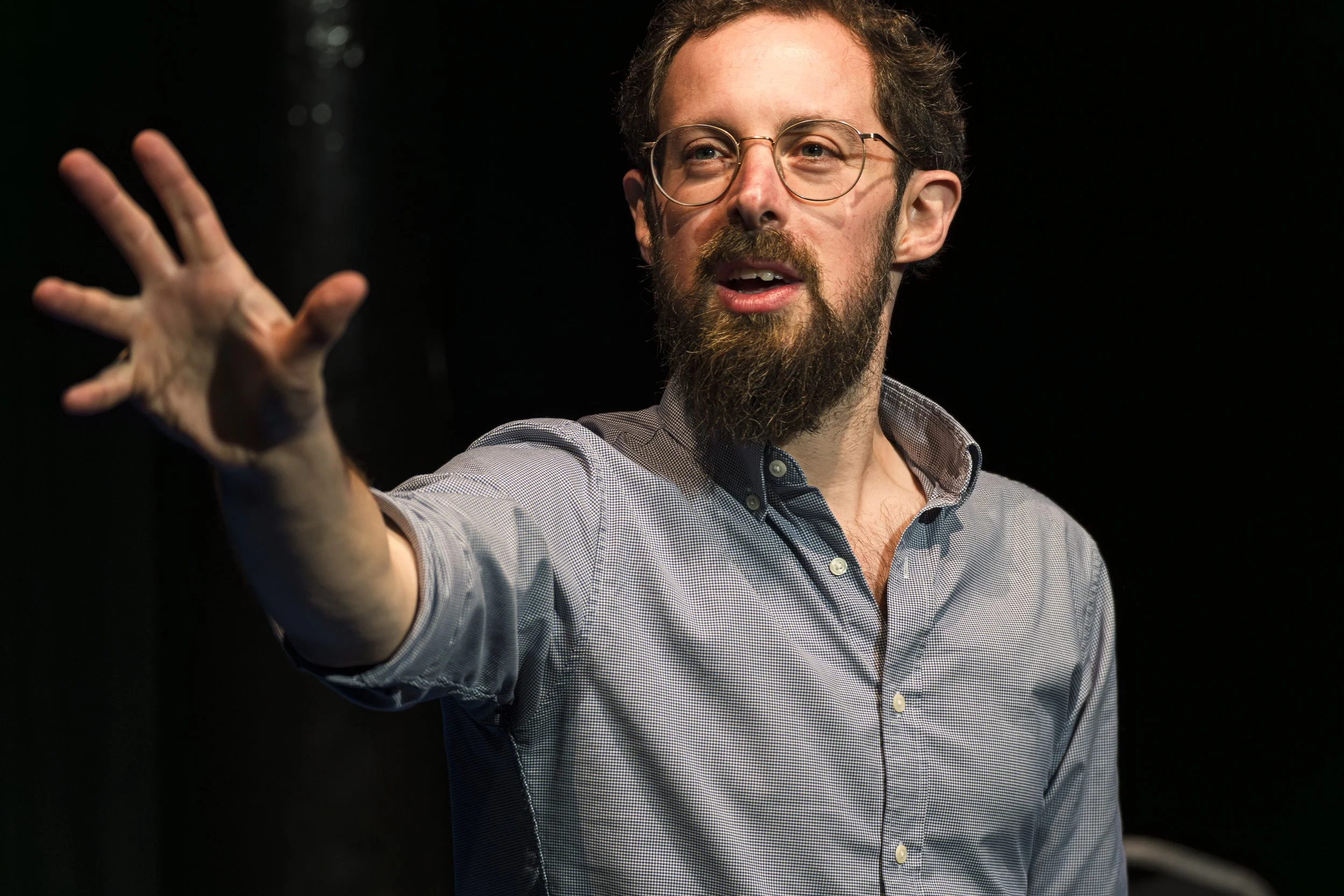A man with glasses, a beard, and a checkered shirt speaking while pointing with his hand on a dark background.