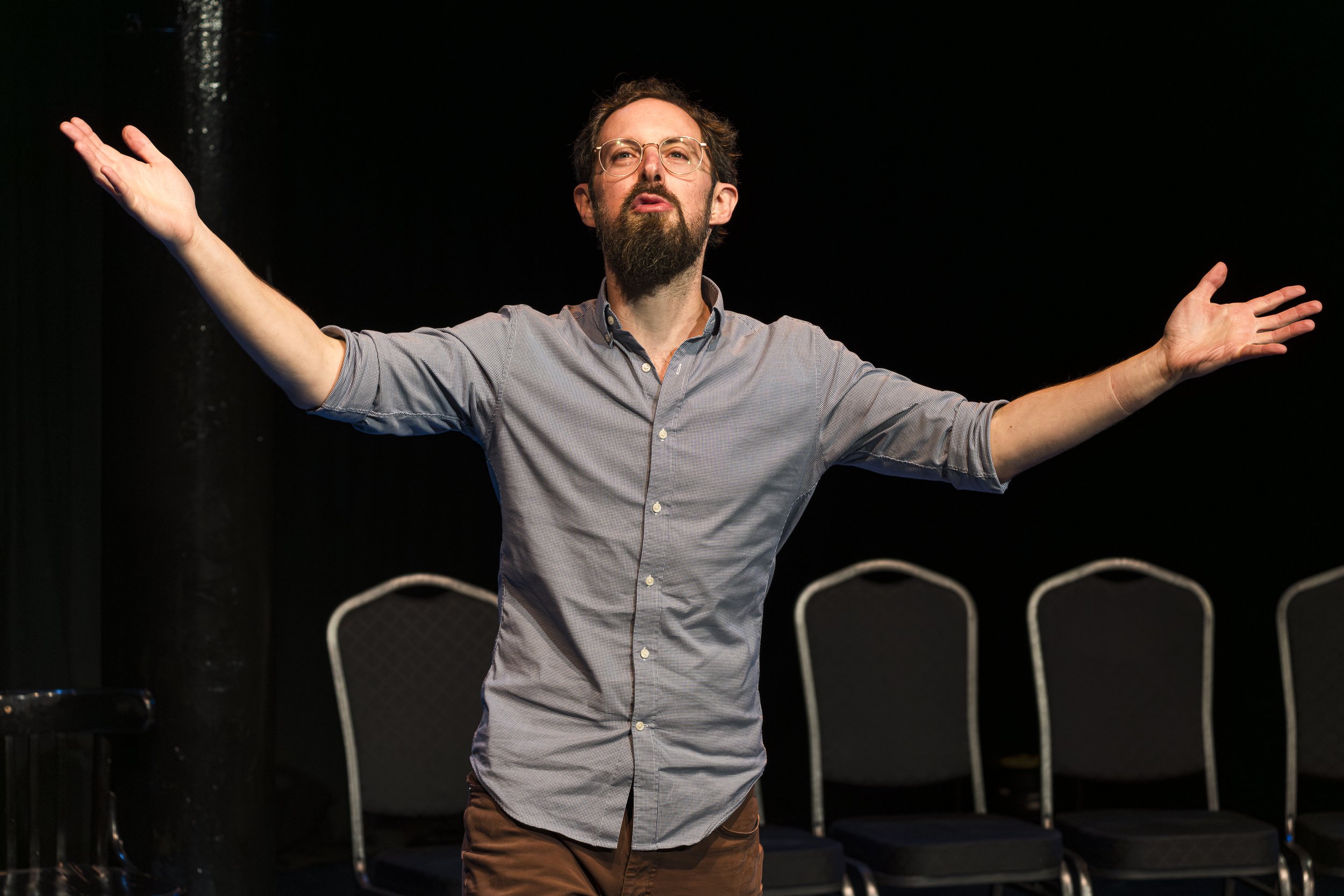 Man with beard and glasses standing with arms outstretched on stage, black background, empty chairs behind him.