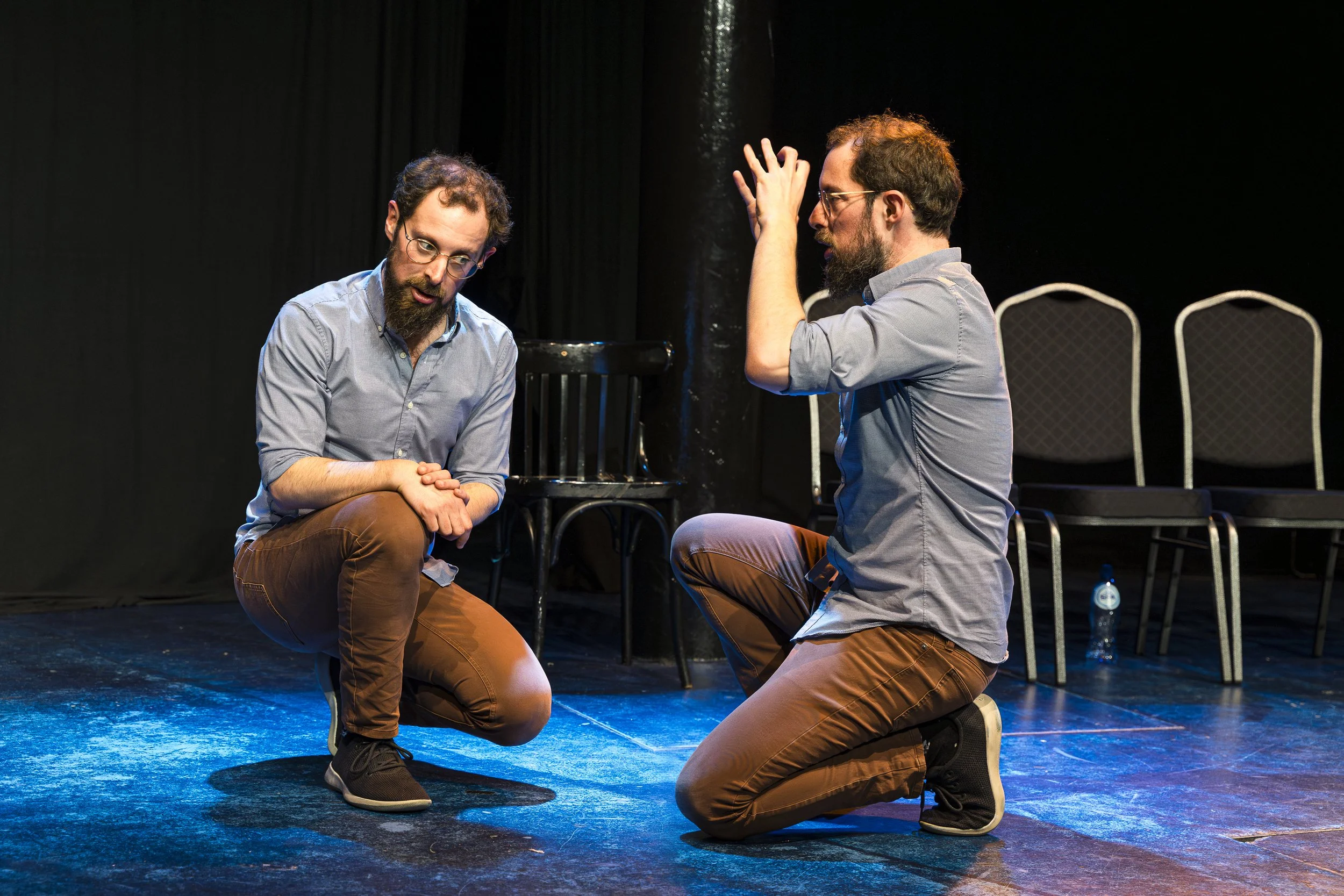 Two men with glasses and beards, wearing gray shirts and brown pants, are performing a scene on a black stage, with chairs and a water bottle in the background. One man is kneeling with hands clasped, and the other is kneeling with hands raised.