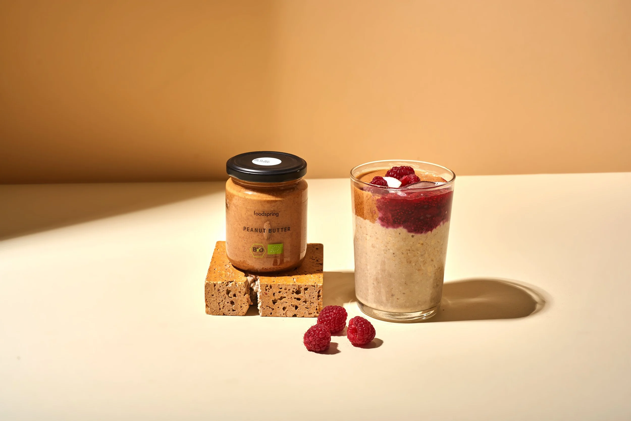 A jar of organic peanut butter, a glass of oatmeal topped with raspberries, and three raspberries on a light-colored surface with a beige background.