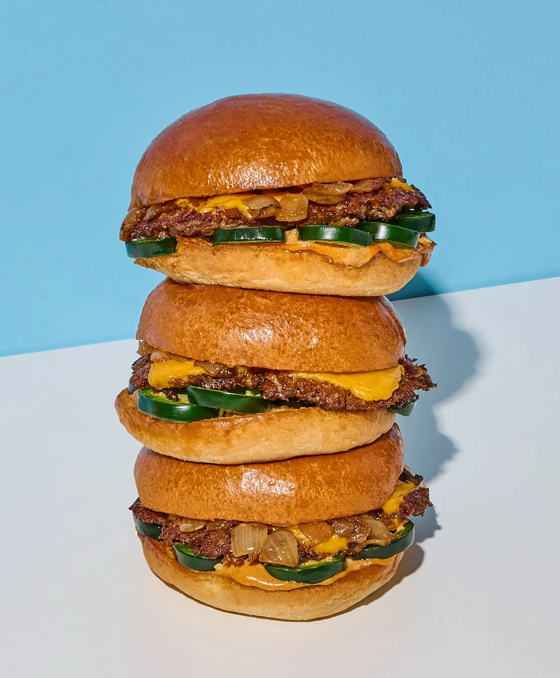 Stack of three cheeseburgers with pickles, onions, and melted cheese on a white and blue background.