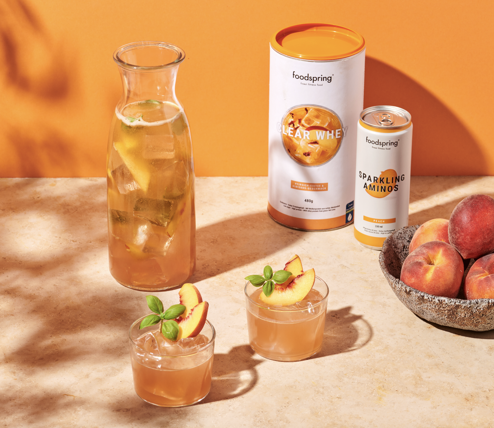 A glass pitcher and two glasses of peach flavored beverage with peach slices and basil garnish, a bowl of peaches, peach protein powder, and sparkling amino drink cans on a beige countertop against an orange background.