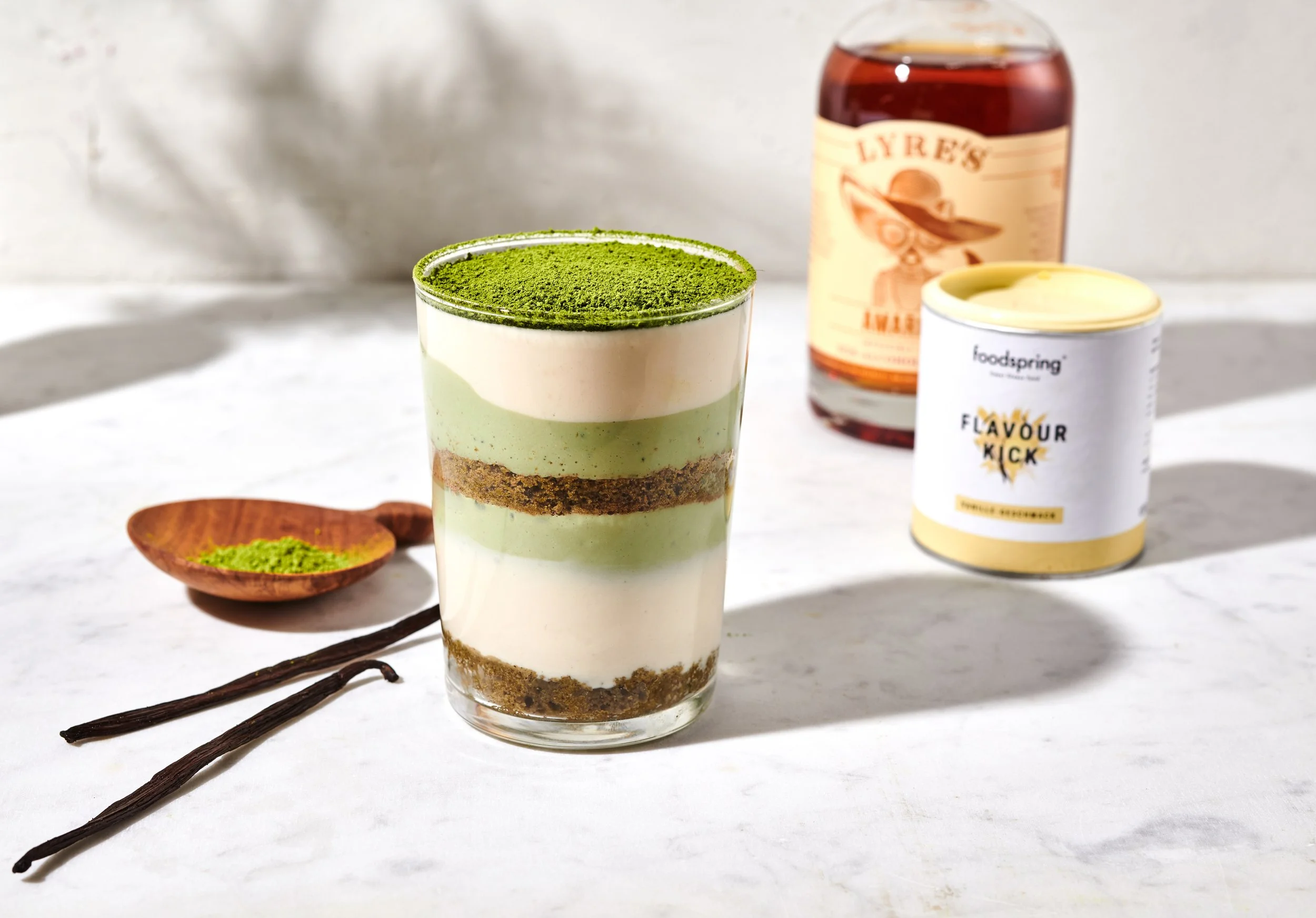 Layered matcha and vanilla dessert in a clear glass, with green powder on top, on a white surface next to vanilla beans, and with a small wooden spoon with matcha powder, with a bottle of Liery's liqueur and a tub of FoodSpring Flavour Kick powdered 