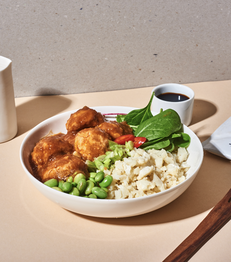 Bowl of Asian-inspired food with meatballs, edamame, mashed potatoes, and greens, accompanied by a small cup of soy sauce or dipping sauce.