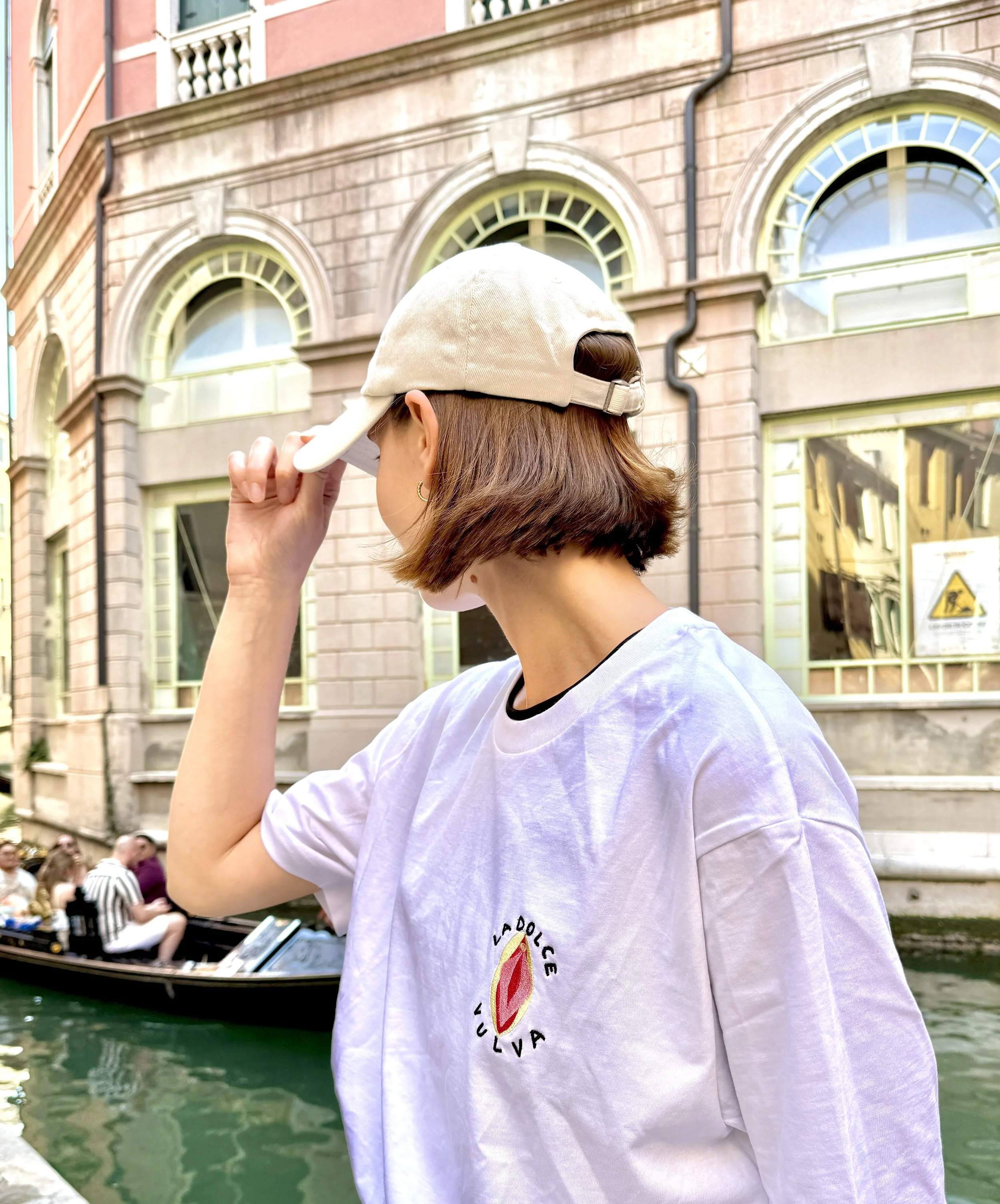 A woman wears a beige cap and a white T-shirt with a colorful logo on the chest, standing near a canal and looking away, with historic pink and beige buildings and a boat with people in the background.
