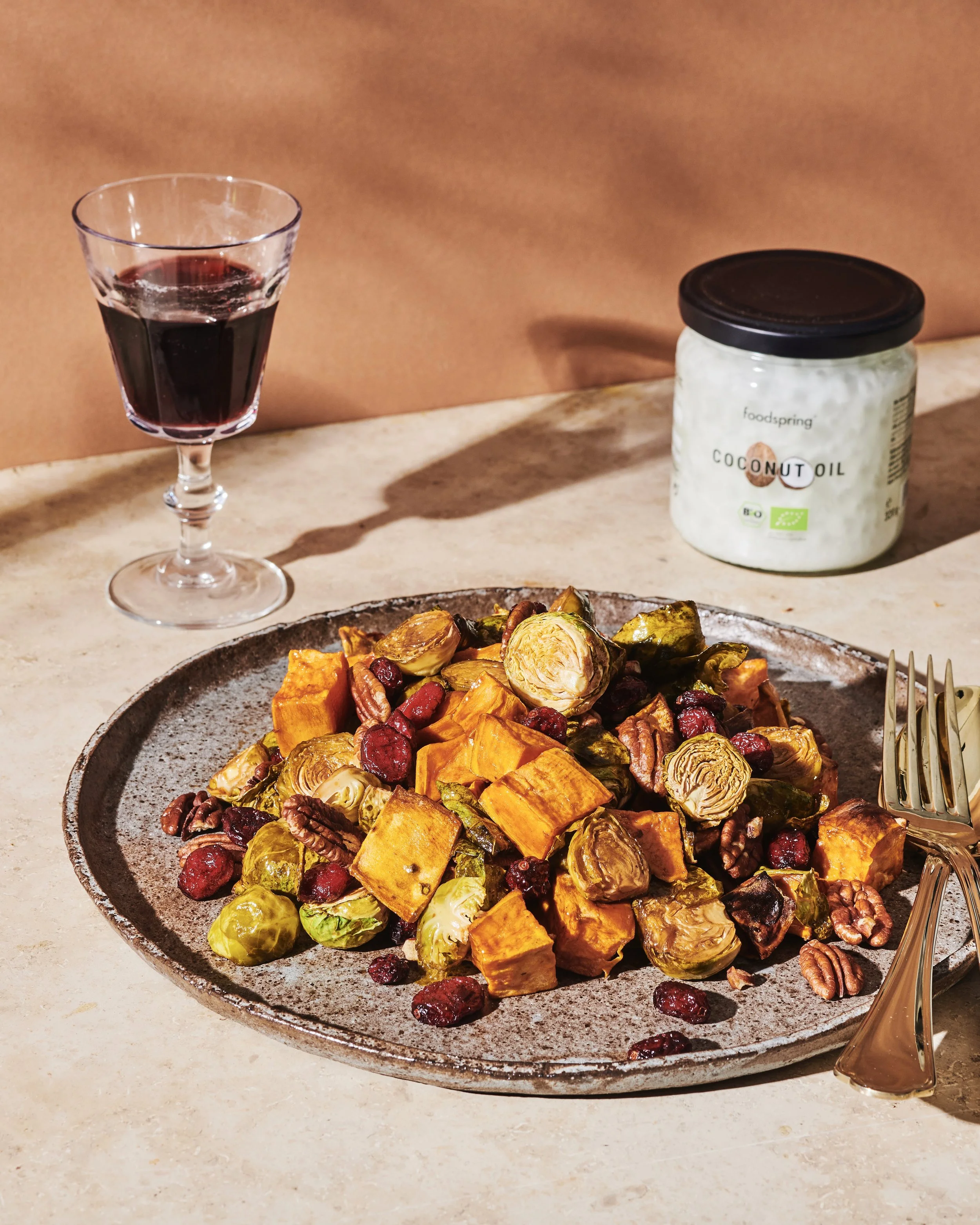 A plate of roasted vegetables includes sweet potatoes, brussels sprouts, pecans, and cranberries on a table. A fork is on the right side of the plate. There is a glass of red wine to the left and a jar of coconut oil behind the plate.