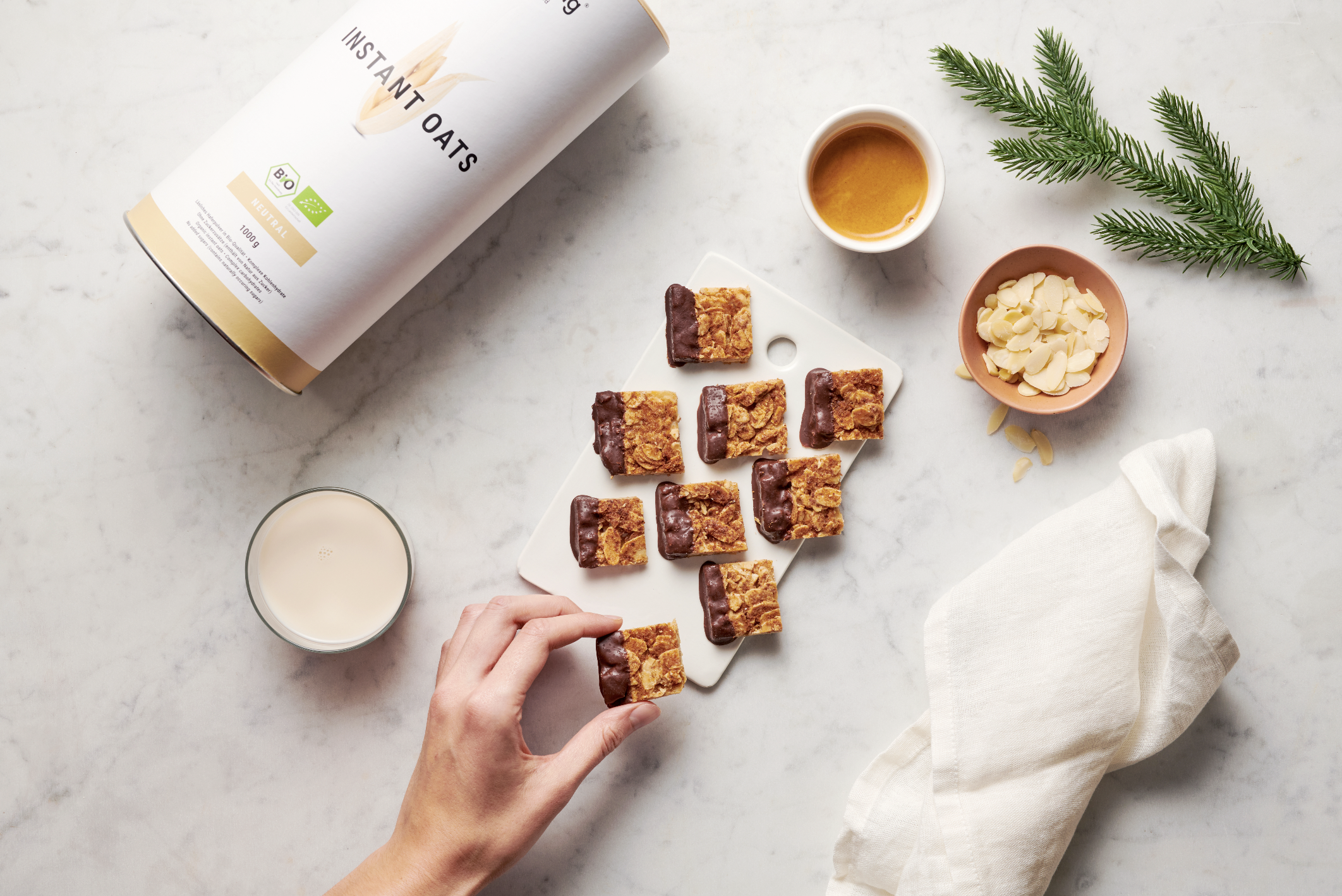 Hand reaching to pick a piece of granola bar on a white marble surface, surrounded by a cup of tea, a glass of milk, a small dish of sliced almonds, a bowl of honey, and a jar of instant oats. A green pine branch is on the top right corner.
