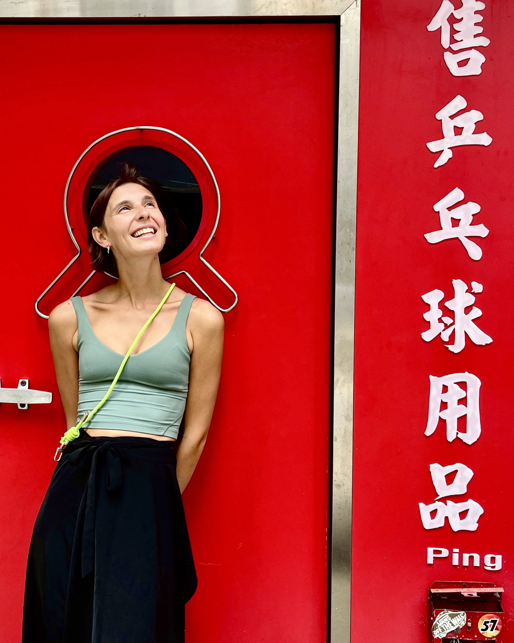 A woman standing in front of a red door with a circular window, smiling and looking up. There are Japanese characters and the word 'Ping' on the right side of the door.