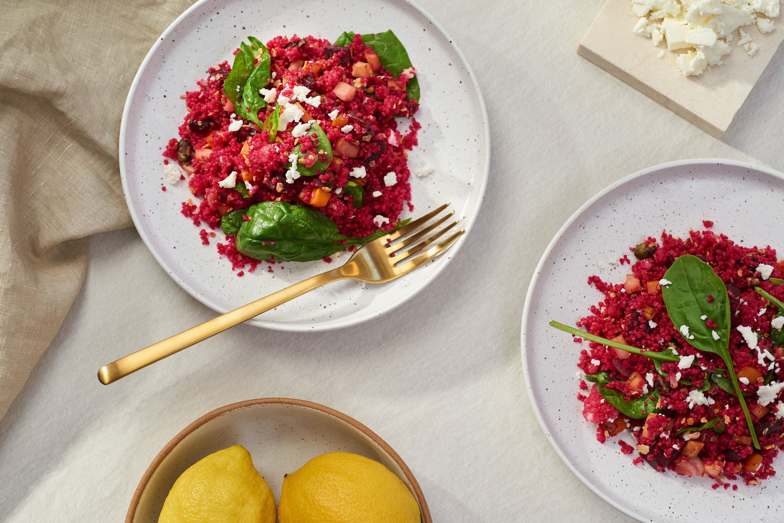 Two white plates of beet salad with greens and crumbled white cheese, and a small bowl of lemons on a table.