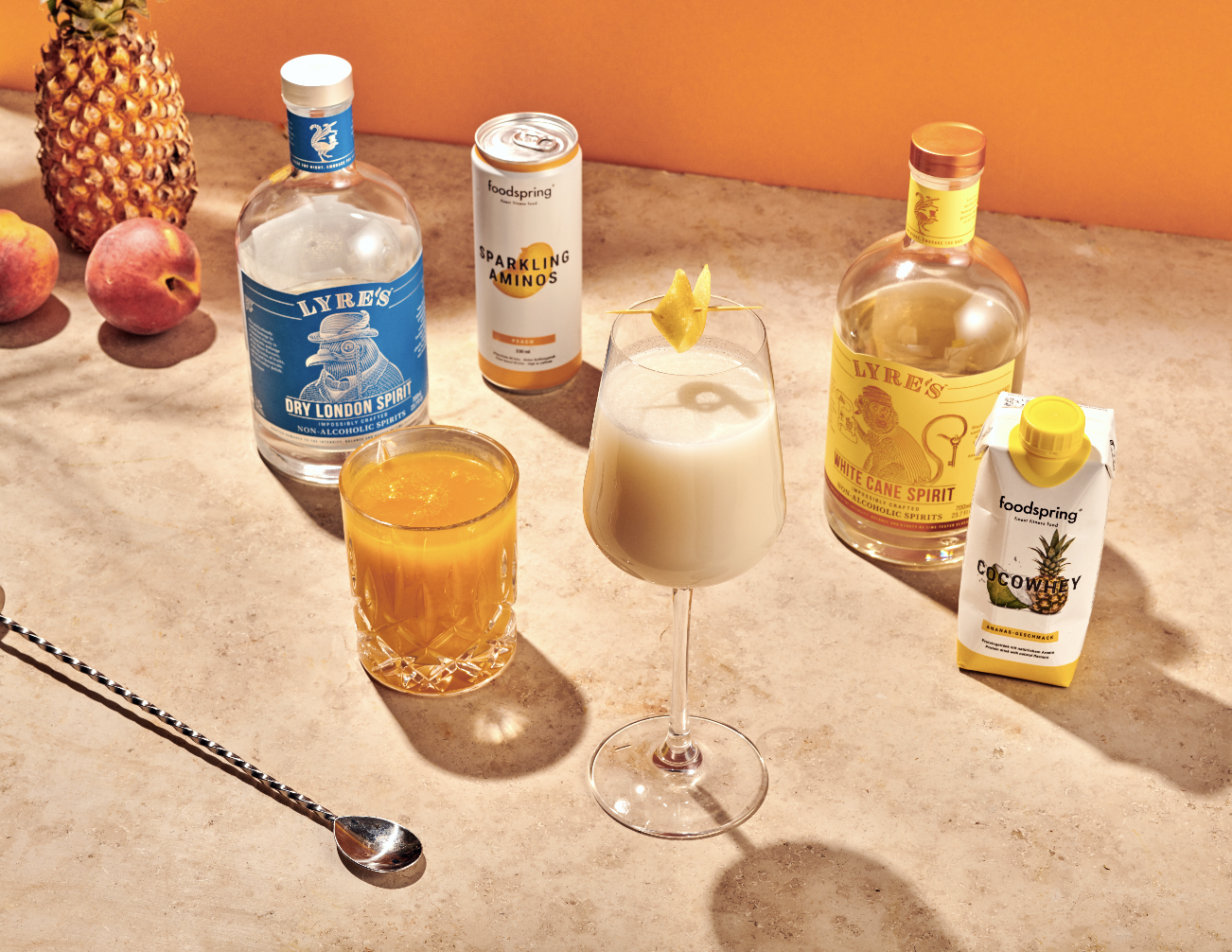 A scene featuring two bottles of spirits, one labeled 'Dry London Spirit' and the other 'White Cane Spirit,' along with a can of Sparkling Aminos, a carton of coconut pineapple juice, a glass of an orange-colored drink, a white cocktail with a pineapple garnish, and a spoon, all arranged on a countertop with some fruit in the background.