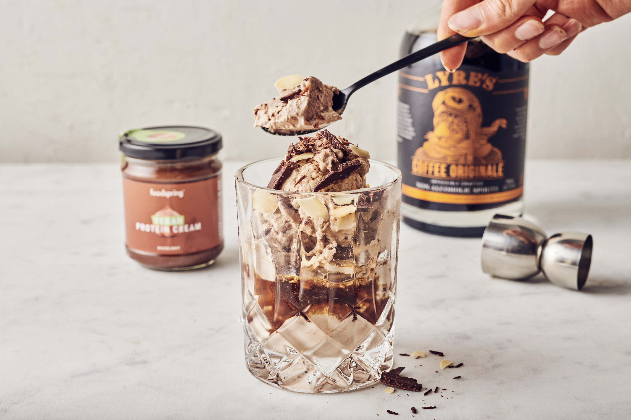 A hand holding a spoonful of chocolate ice cream over a glass filled with ice cream and chocolate syrup. In the background, there is a jar of vegan protein cream, a container of coffee, and a metal jigger on a white surface.