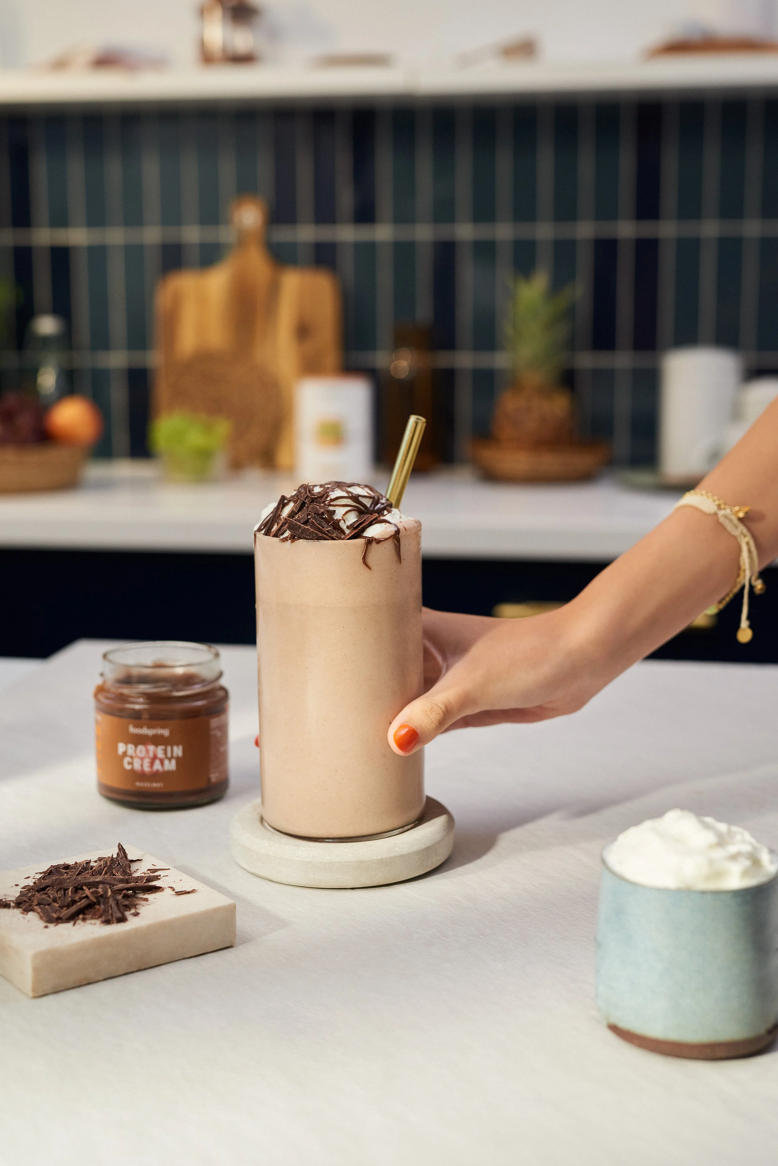 A hand holding a tall glass of chocolate protein shake topped with whipped cream and chocolate shavings, with a straw. There is a jar of chocolate protein cream and a bowl of whipped cream on the table.